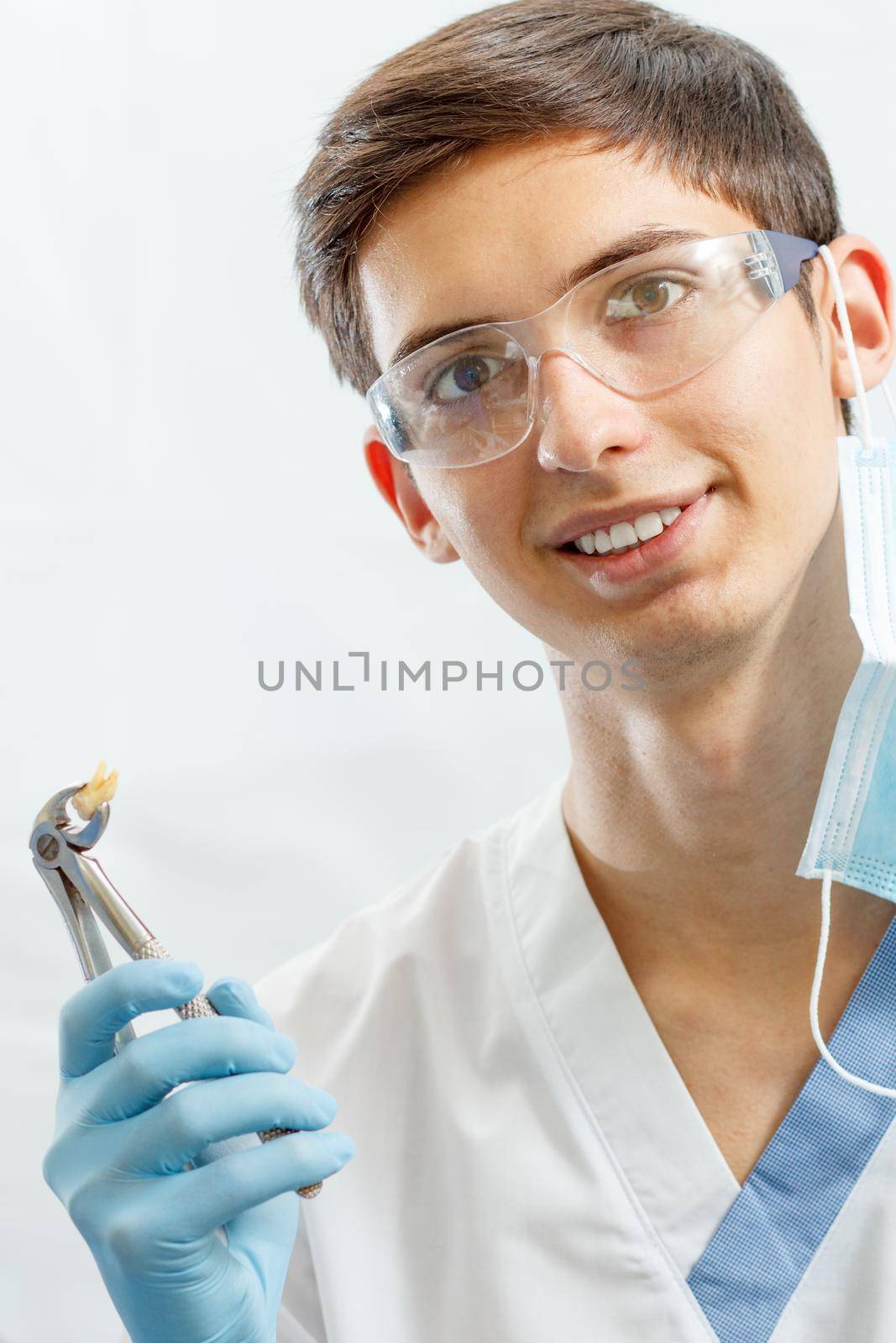 Portrait of handsome male dentist with stainless steel dental tongs or pliers and extracted lower tooth in it. Doctor wearing white uniform, glasses and blue gloves