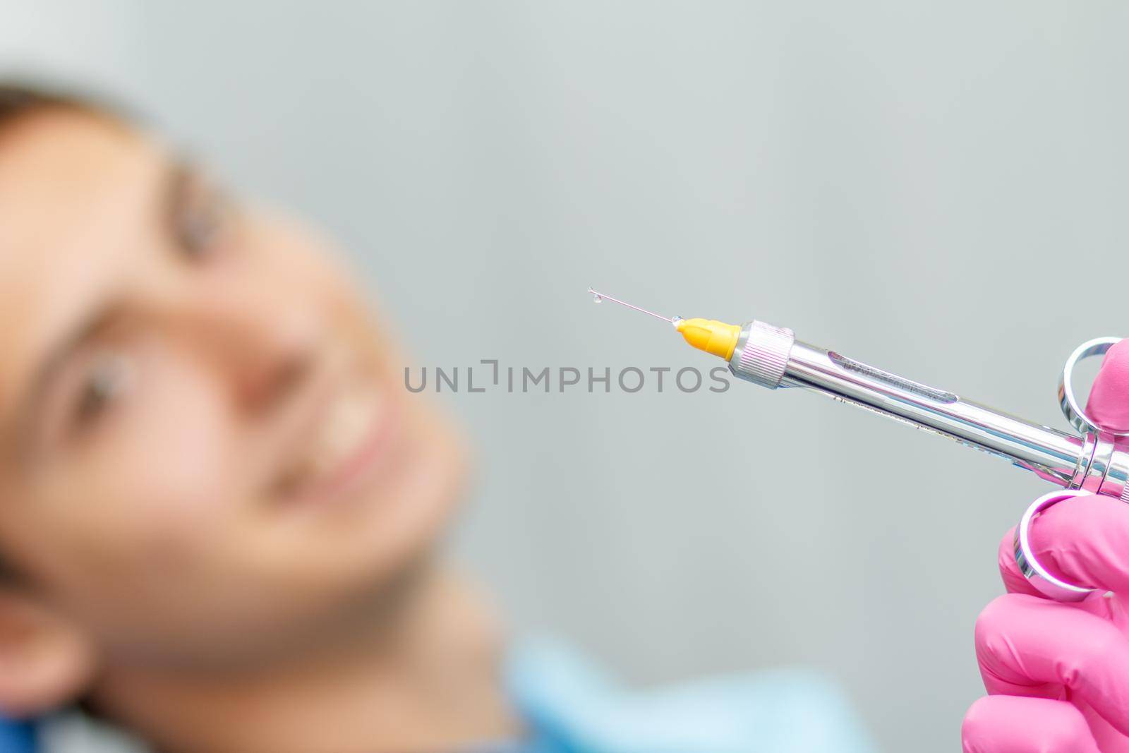 Doctor wearing pink gloves is holding syringe with anesthetic and needle with young man smiling on the background by mvg6894