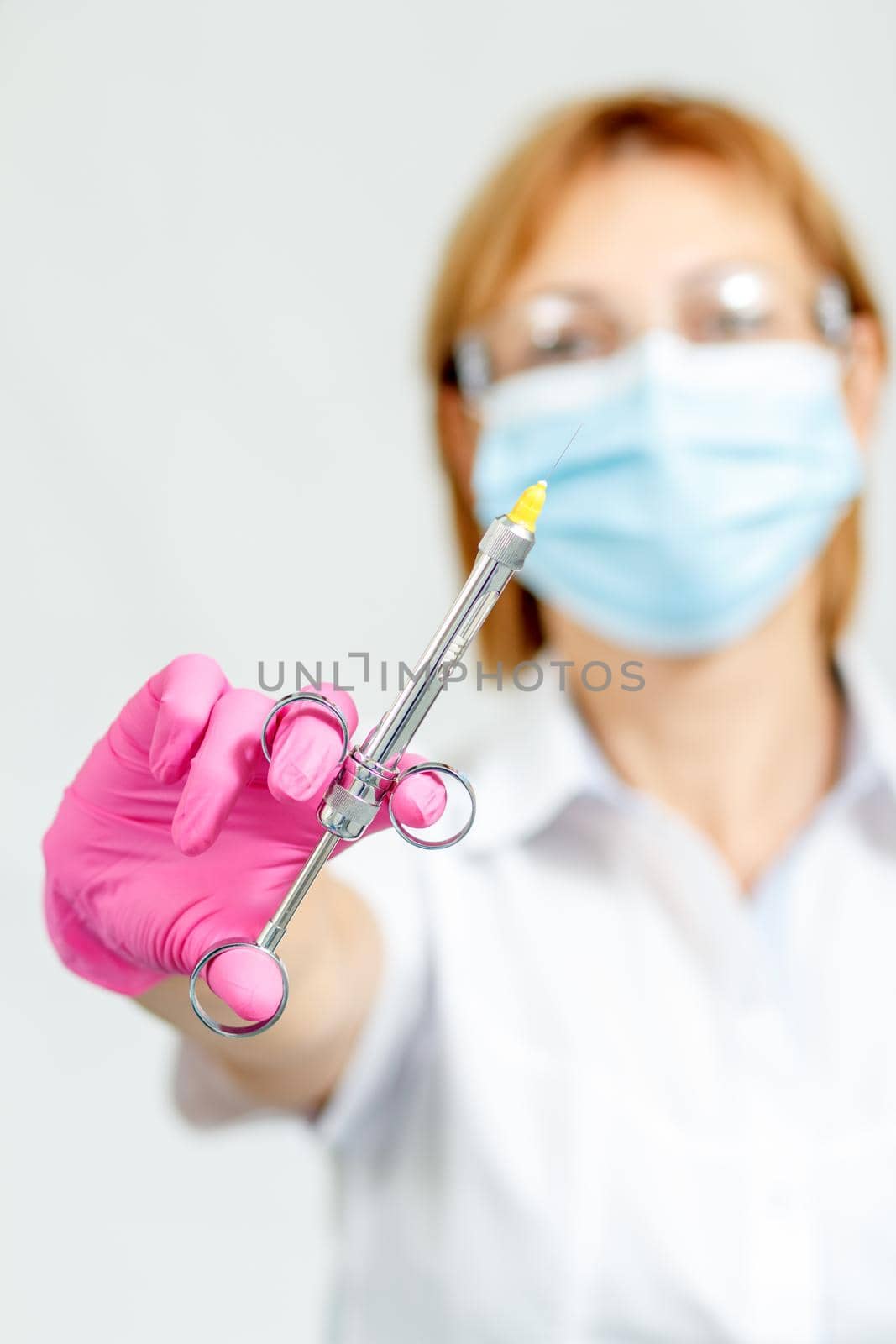 Female doctor in mask, glasses, pink glove holding syringe with anesthetic and needle. Selective focus on syringe