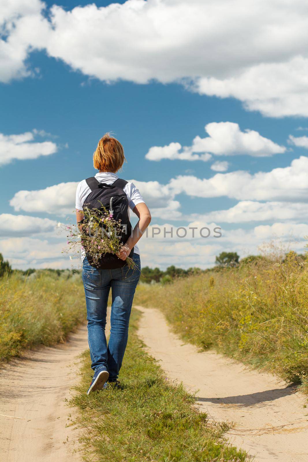 Young lady walking on a rural road. Hiker with backpack walking outdoors in nature. Traveler walking along country trail outdoor. Healthy active lifestyle
