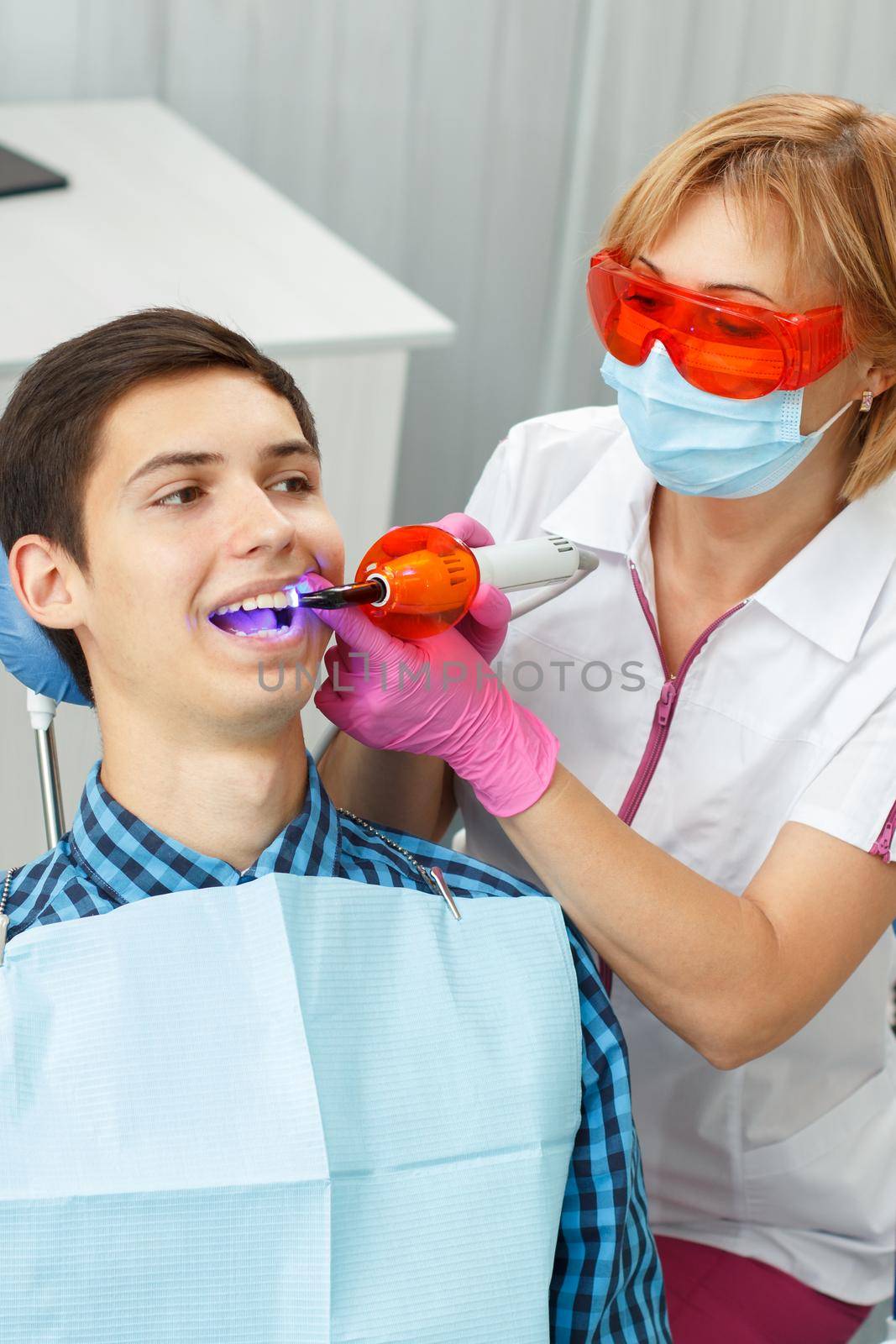 Beautiful woman dentist treating a patient teeth in dental office. Doctor wearing glasses, mask, white uniform and pink gloves. Dentistry
