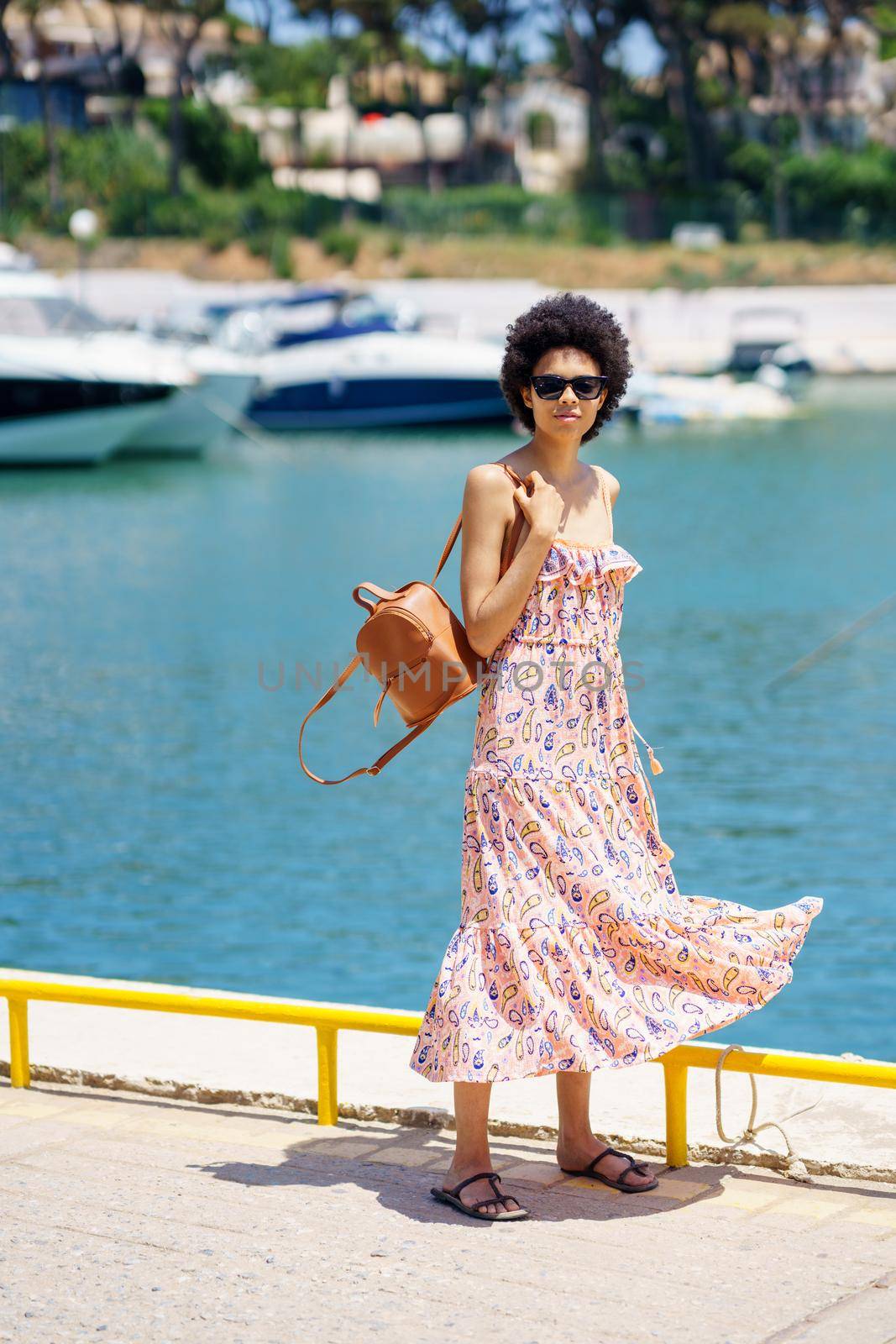 Full body ethnic female tourist in long dress fluttering on wind carrying bag on shoulder, and looking at camera while standing on embankment on sunny summer day in city harbor