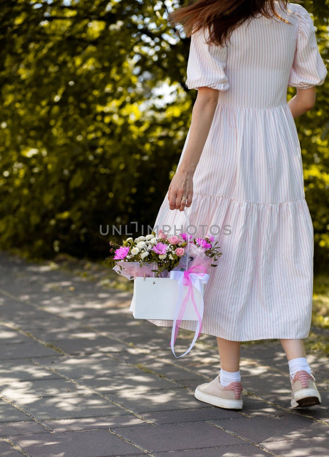 A beautiful bouquet of flowers in a box in the hands of a beautiful girl who walks along the street on a sunny day. Girl in a dress, glasses and sneakers. Focus on the background of flowers.