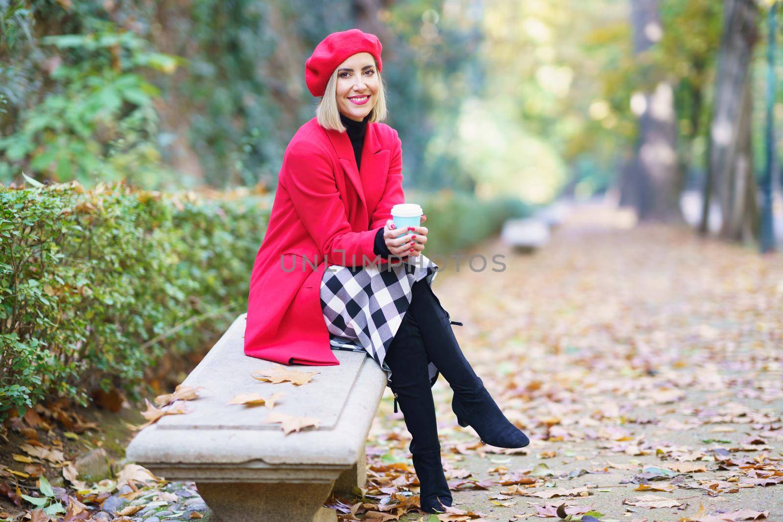 Full body of elegant female, sitting on bench with crossed legs and warming hands, with cup of hot takeaway beverage in autumn park and looking at camera