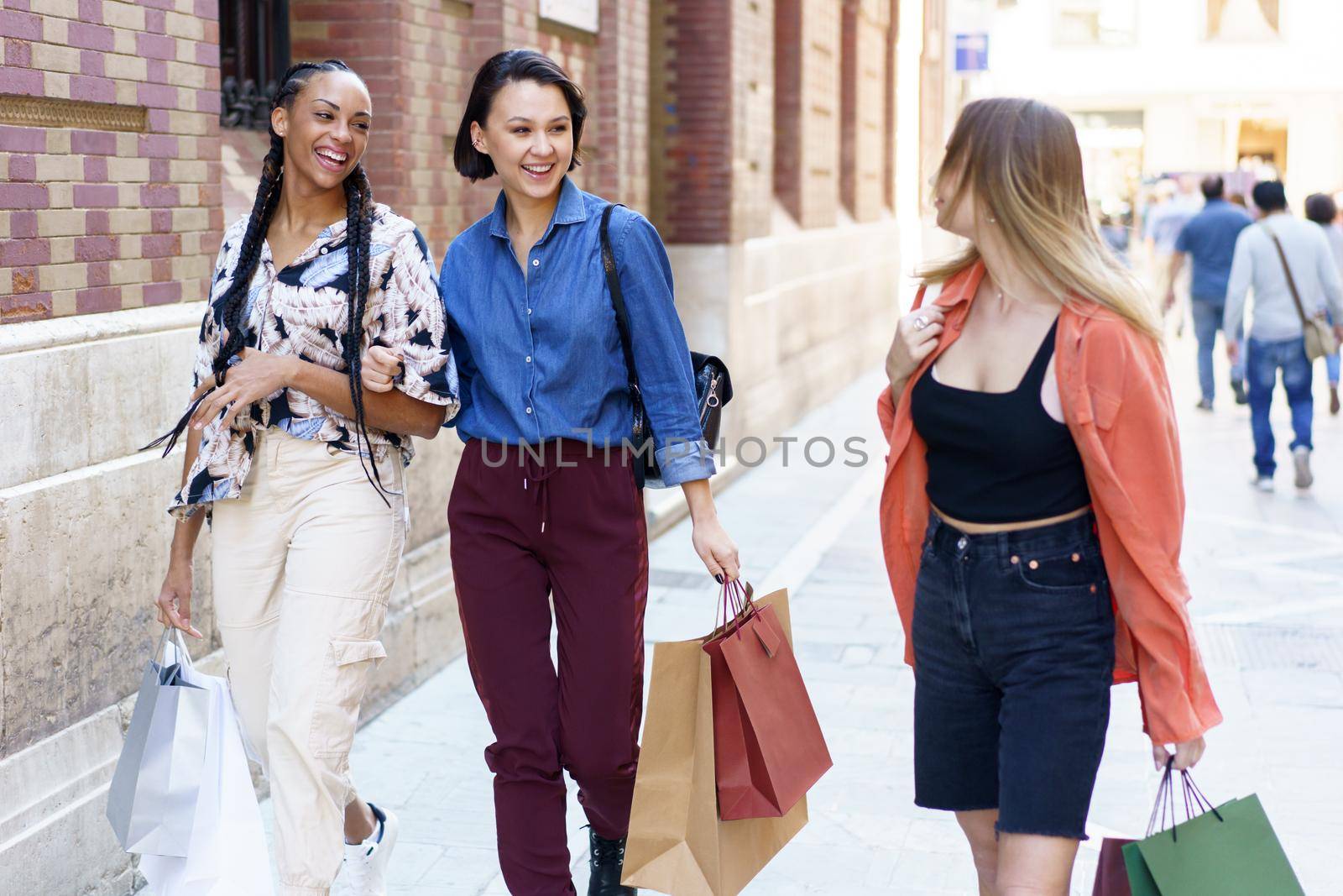 Group of cheerful multiracial female friends with shopping bags of purchases looking at each other while strolling on sidewalk near building in city