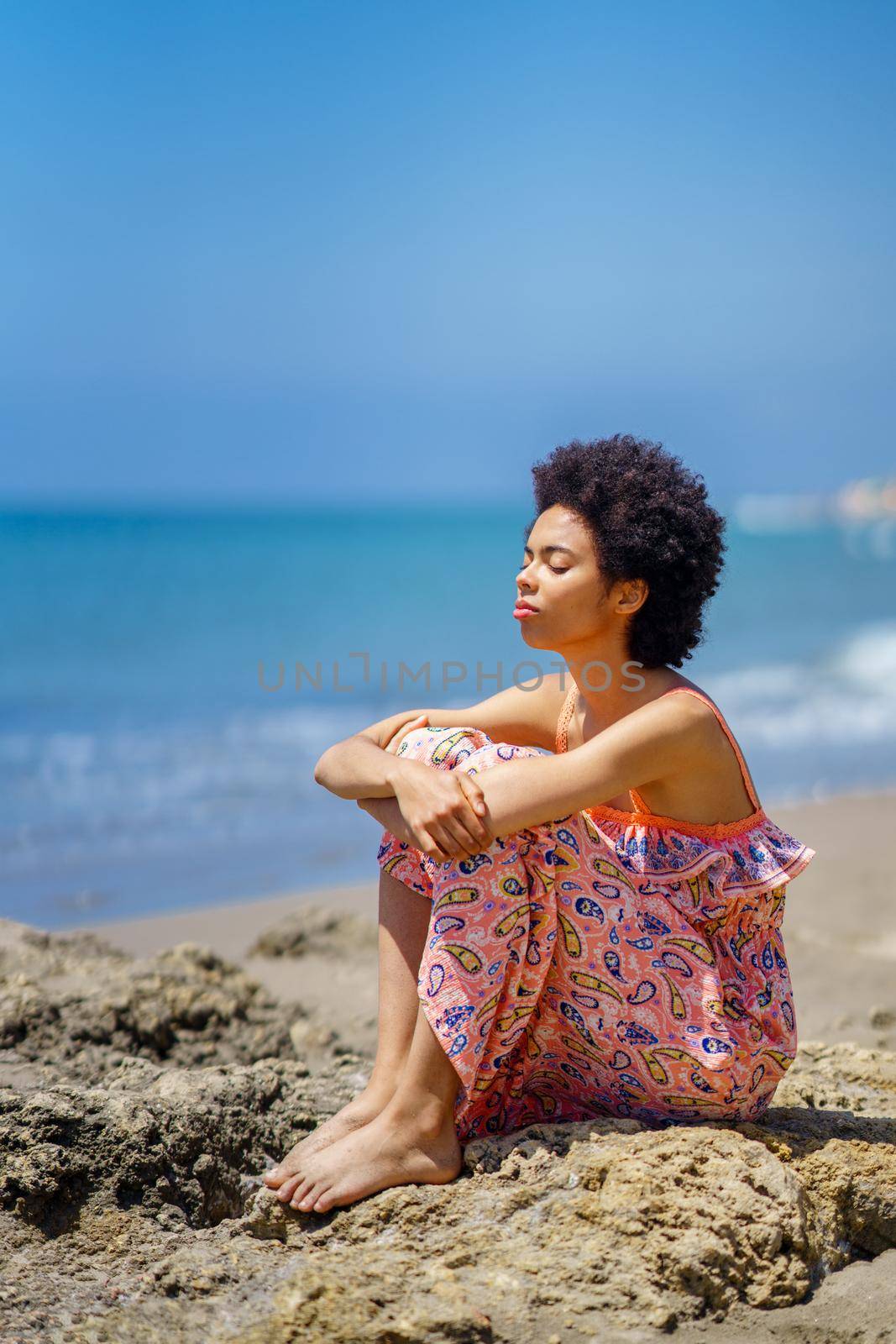 Tranquil ethnic female in maxi dress with curly hair and closed eyes, embracing knees while sitting on stones against waving sea and blue sky on summer weekend day on beach