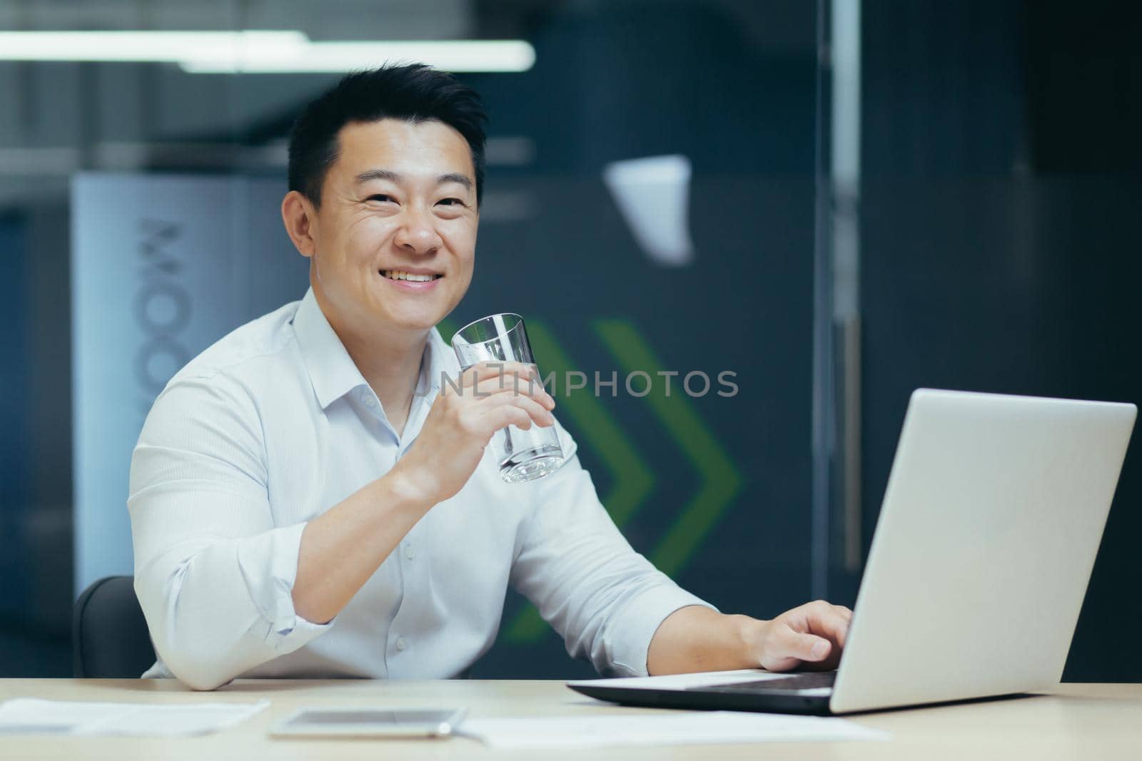 Asian businessman in the office drinking clean filtered water, portrait of a man smiling and looking at the camera holding a glass of water, a man at work with a laptop