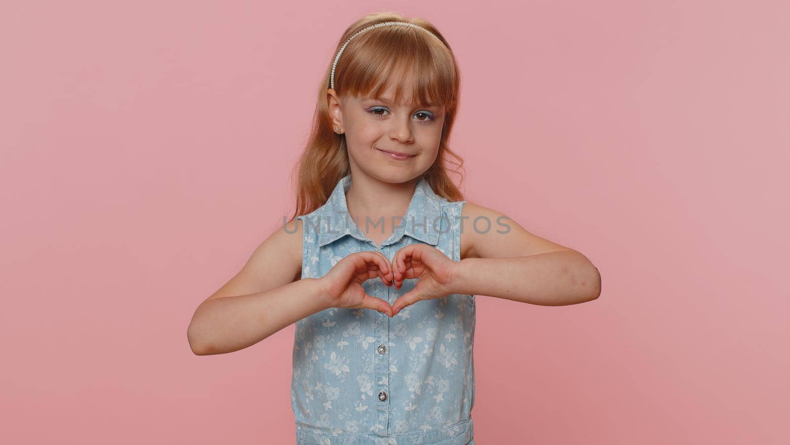 I love you. Smiling young preteen child girl kid makes heart gesture demonstrates love sign expresses good feelings and sympathy. Little toddler children isolated alone on studio pink background