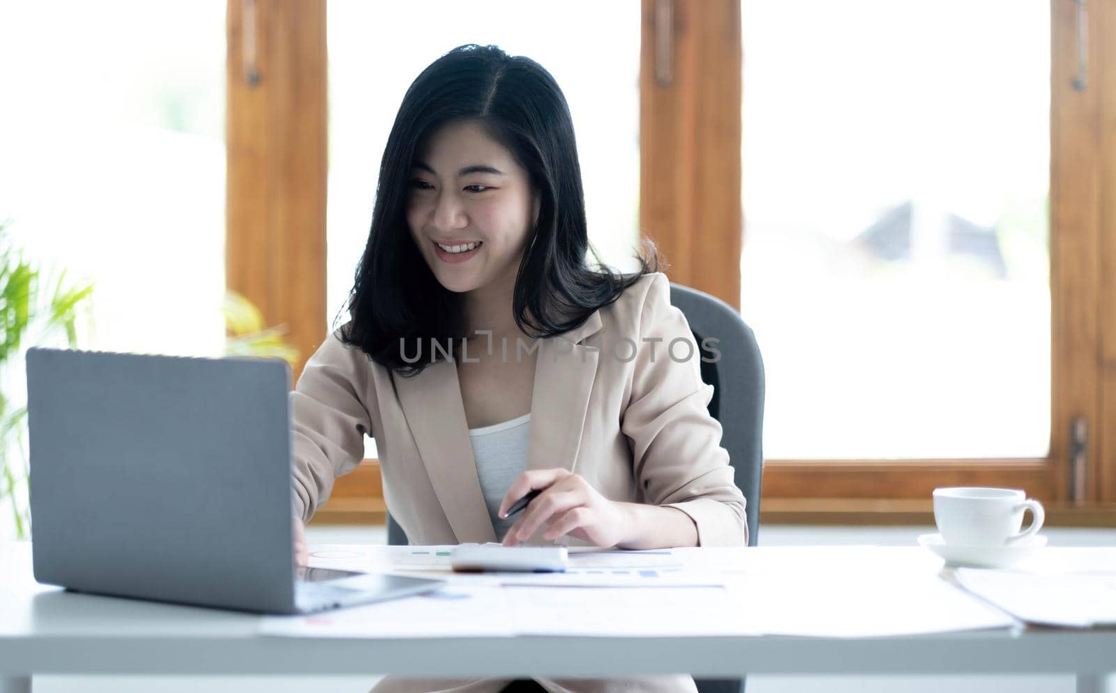 Beautiful Asian businesswoman analyzes charts using laptop calculator at the office. by wichayada