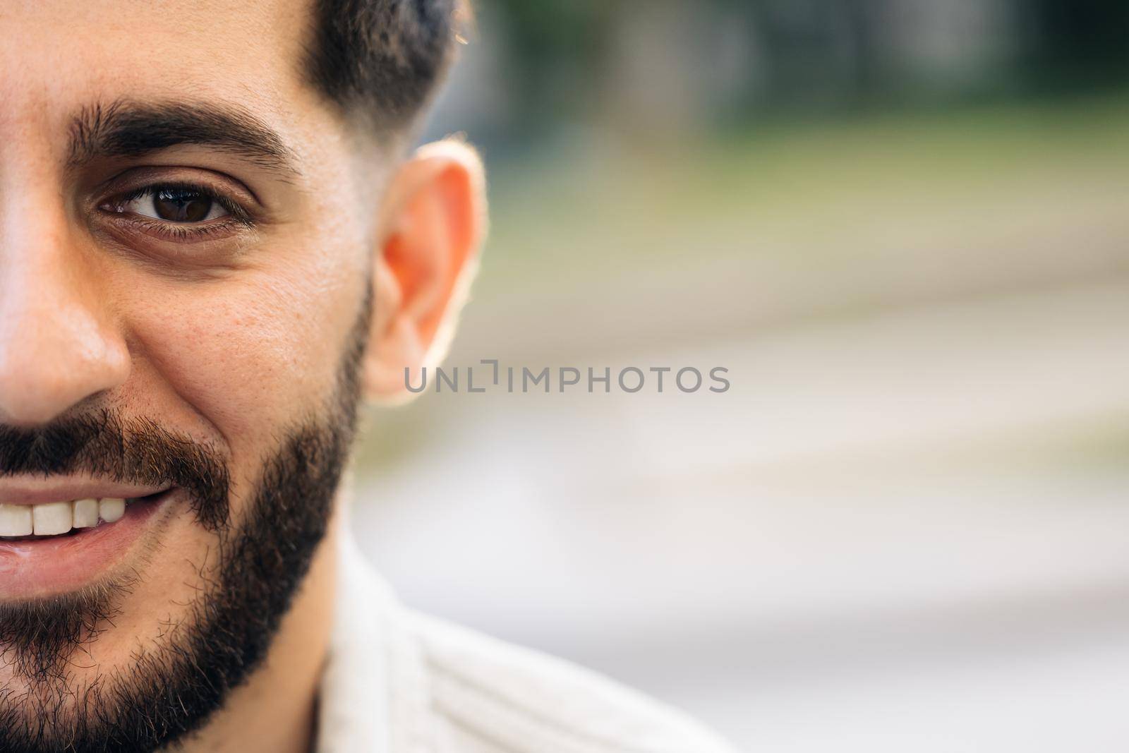 Half face of bearded man with brown eye while standing outdoors. Portrait of male looking to camera and smiling, with space for text. Sensual guy with trendy hairstyle looking into camera.