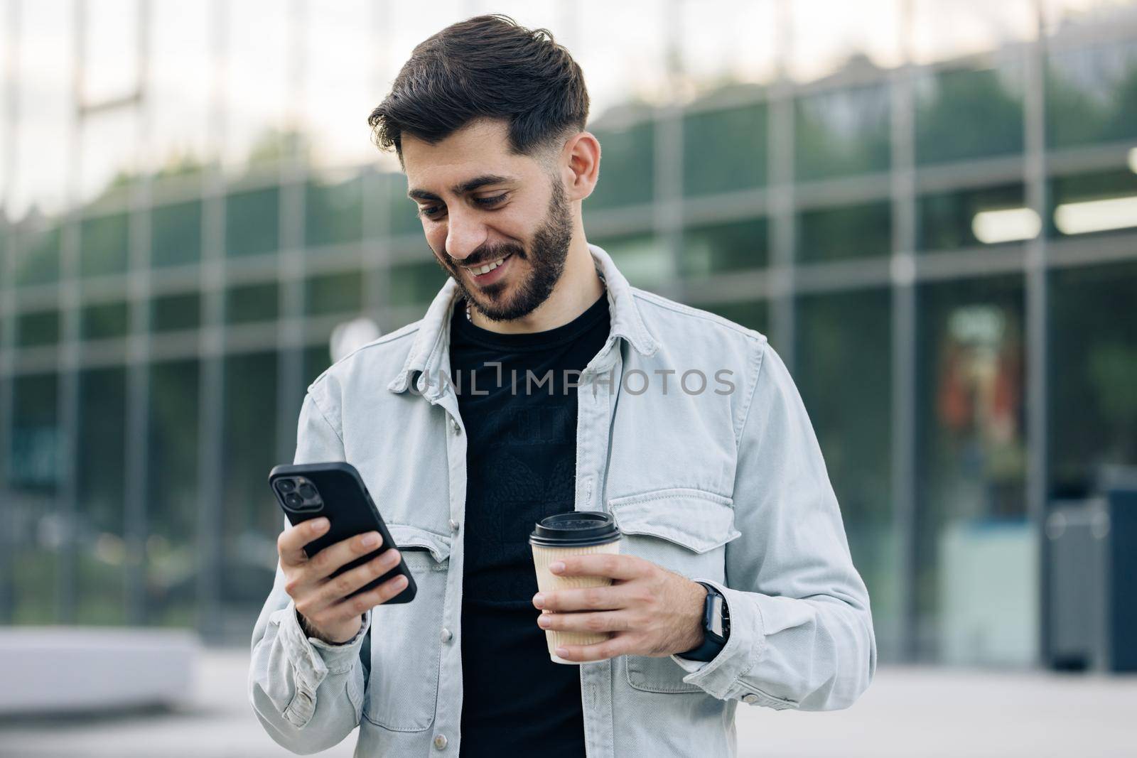 Young elegant bearded man using social media application on smartphone text messages receive news smiling outdoor. People portraits. Technology.