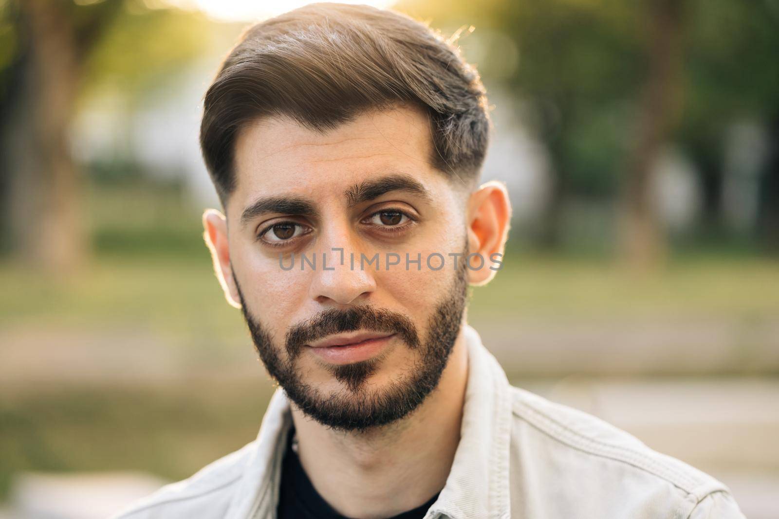 Close up of face young caucasian bearded handsome man looking at camera serious attractive beautiful male stand on street city lifestyle summer guy city outdoors.