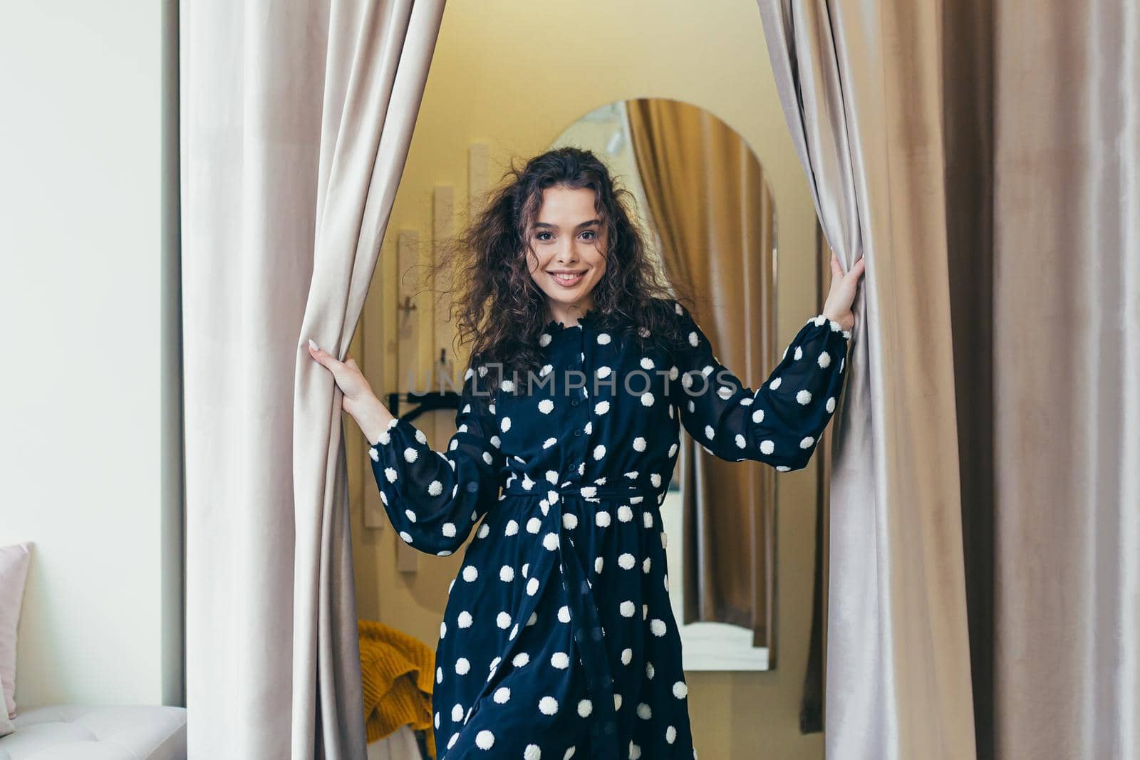 Young beautiful happy woman girl trying on a black dress with white dots near the mirror in the dressing room in a clothing store in the mall. Shows, looks at the camera, poses, brags, wants to buy
