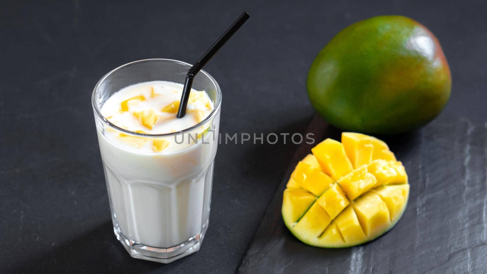 Milk drink on black background with mango. A classic mango milkshake - Lassi. A traditional drink in India from the heat. Top view. Cool mango milkshake on a black background. A drink to quench your thirst in the summer. Indian cuisine classic drink - Lassi. Milk, yogurt with mango chunks