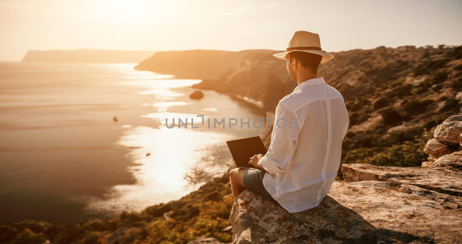 Digital nomad, freelancer. Remote work on vacation. Man in the hat, a businessman with a laptop sits on the rocks by the sea during sunset, makes a business transaction online from a distance. by panophotograph