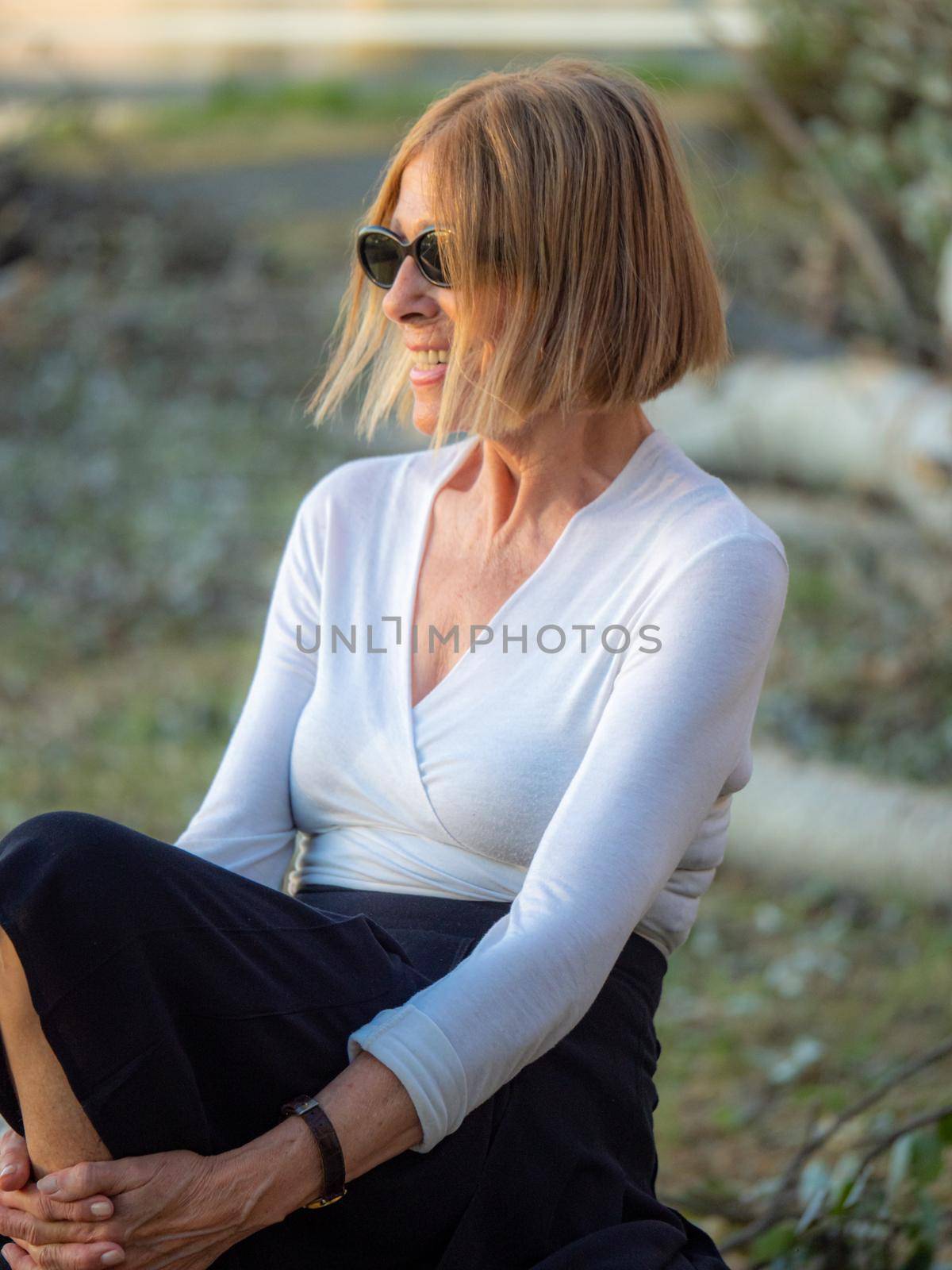 elegant caucasian senior woman wearing sunglasses sitting in a park at dusk by verbano