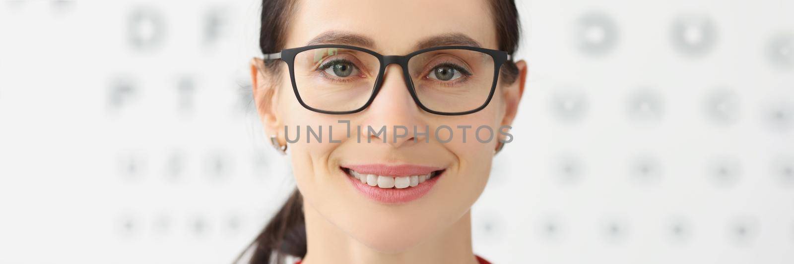 Portrait of smiling female eye doctor with eye test chart on background, female optician in glasses, appointment in eye clinic. Ophthalmologist concept