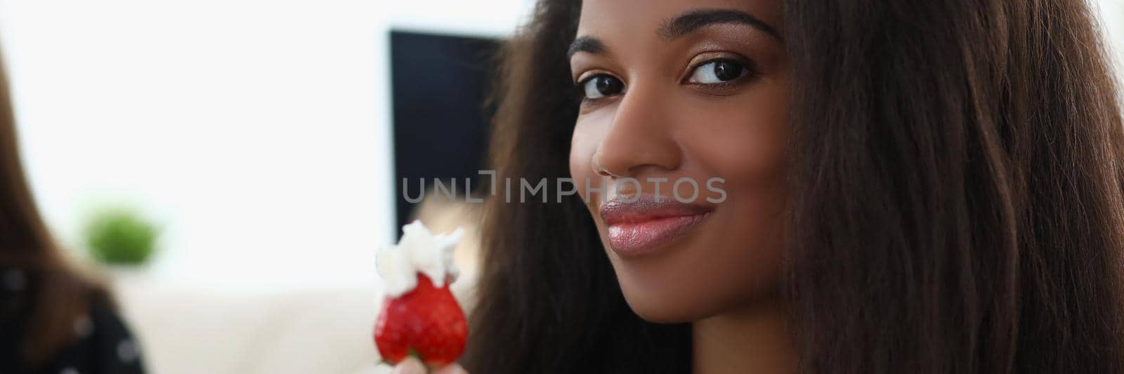 Close-up of latin woman hold fresh strawberry fruit with whipped cream on top. Afro american girl eating berry and smiling. Vitamin, lunch, snack concept