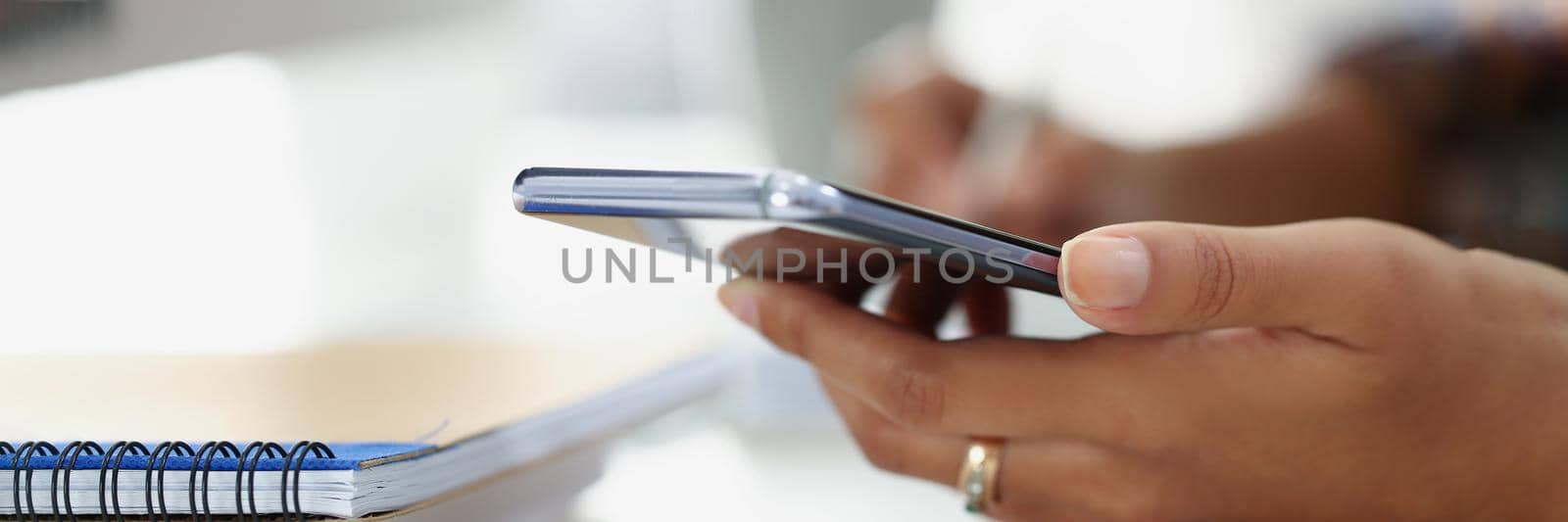 Woman using modern smartphone while sitting at table with spiral notebook in office by kuprevich