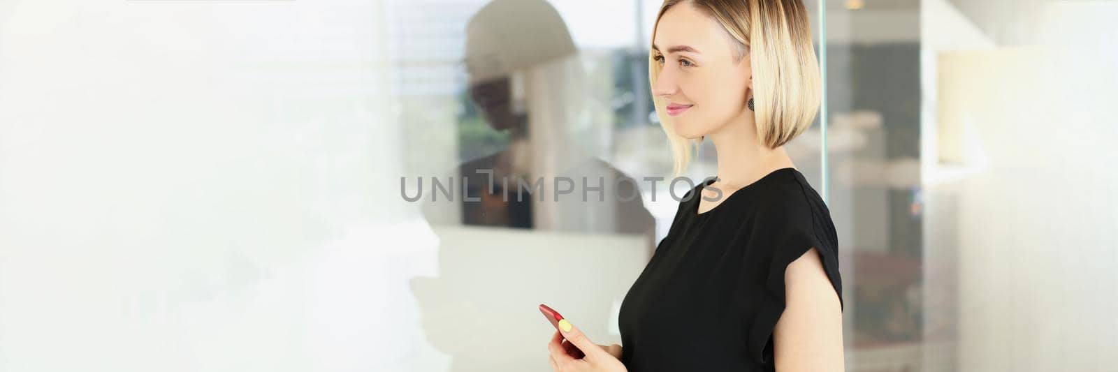Portrait of attractive young blonde businesswoman in stylish black blouse with smartphone in company office. Business, corporation, success, career concept