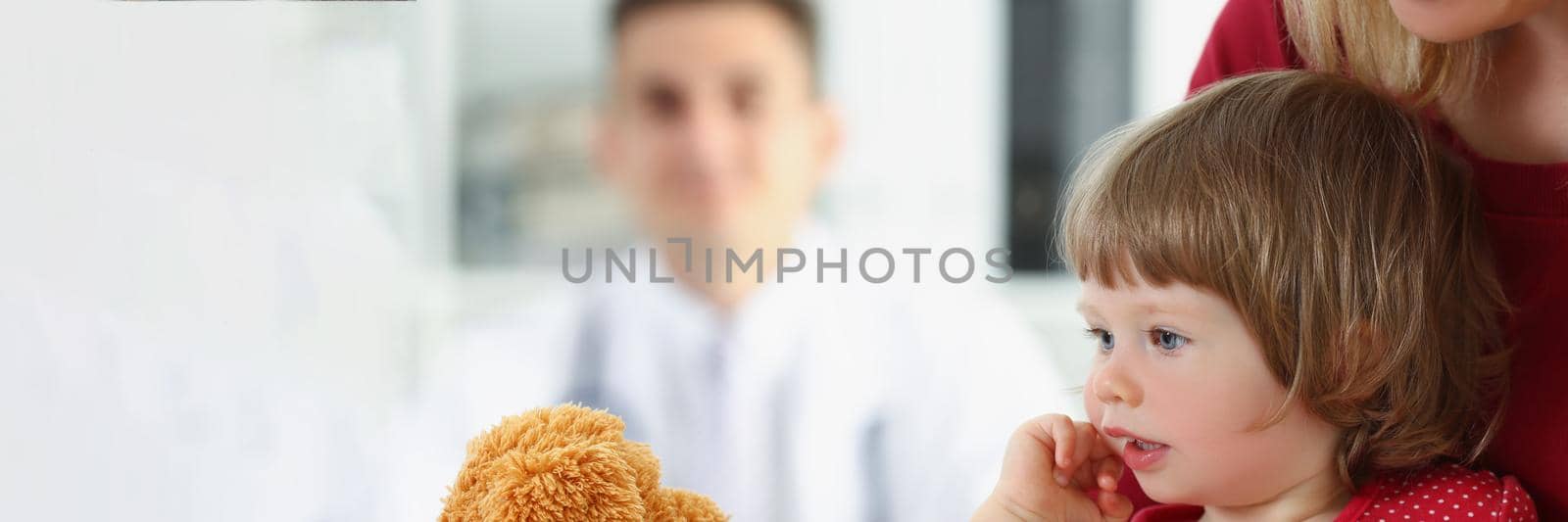 Portrait of little girl with mother play with teddy bear before doctor appointment. Cute child with parent visit family doctor. Hospital, health concept