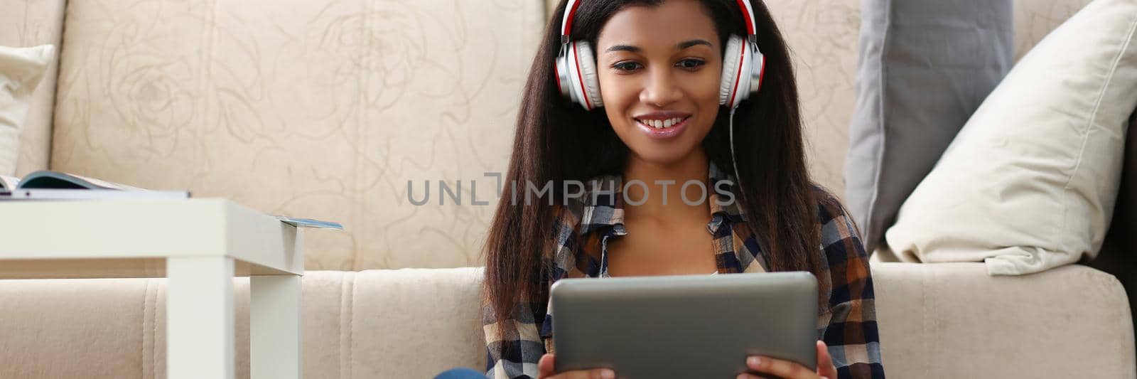 Low angle of latin young woman sit on carpet with tablet and watch show, lady in headset. Spend time at home, enjoy weekend or day off. Leisure concept