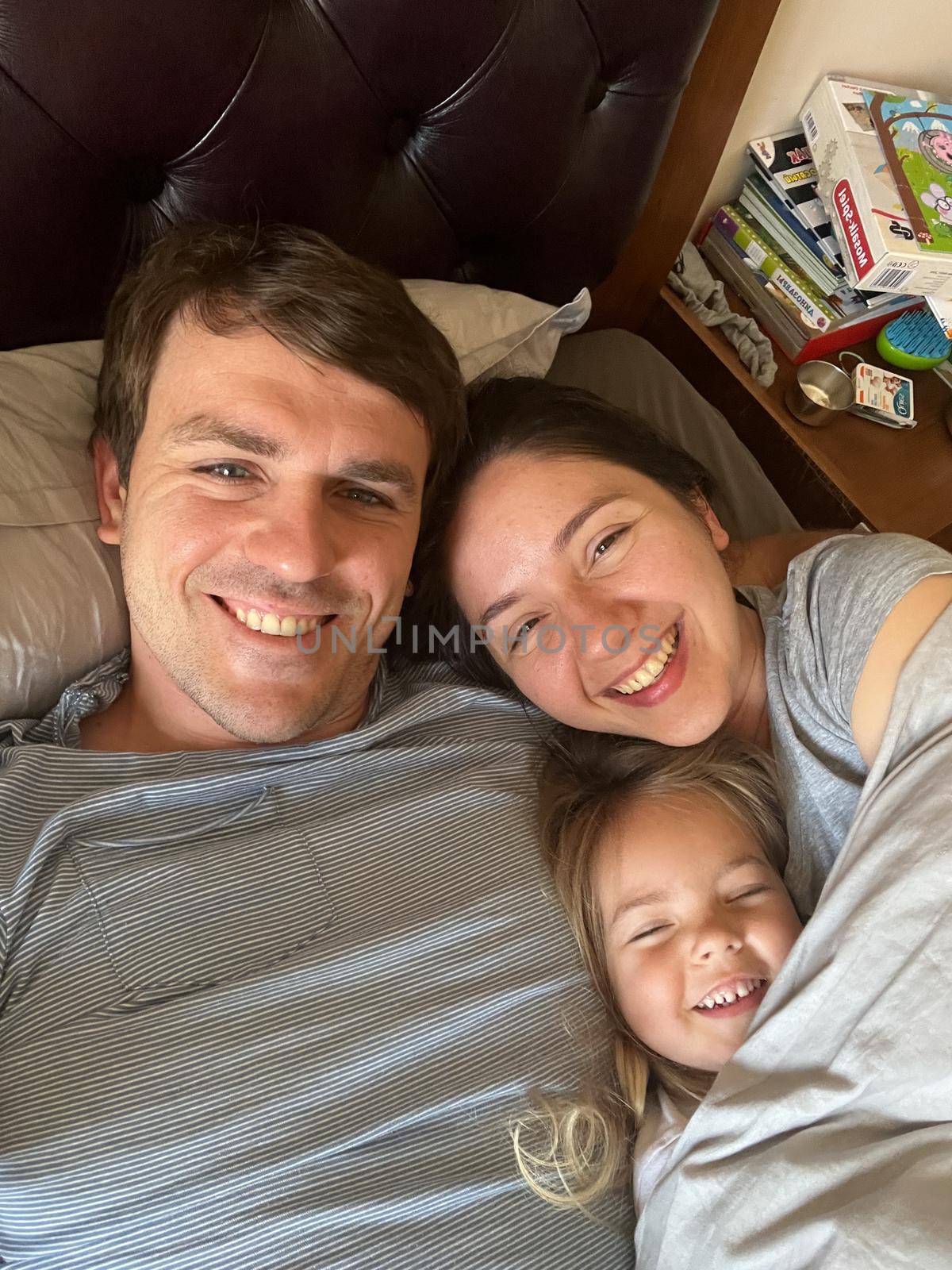 Smiling mom, dad and little daughter lie in bed by Nadtochiy