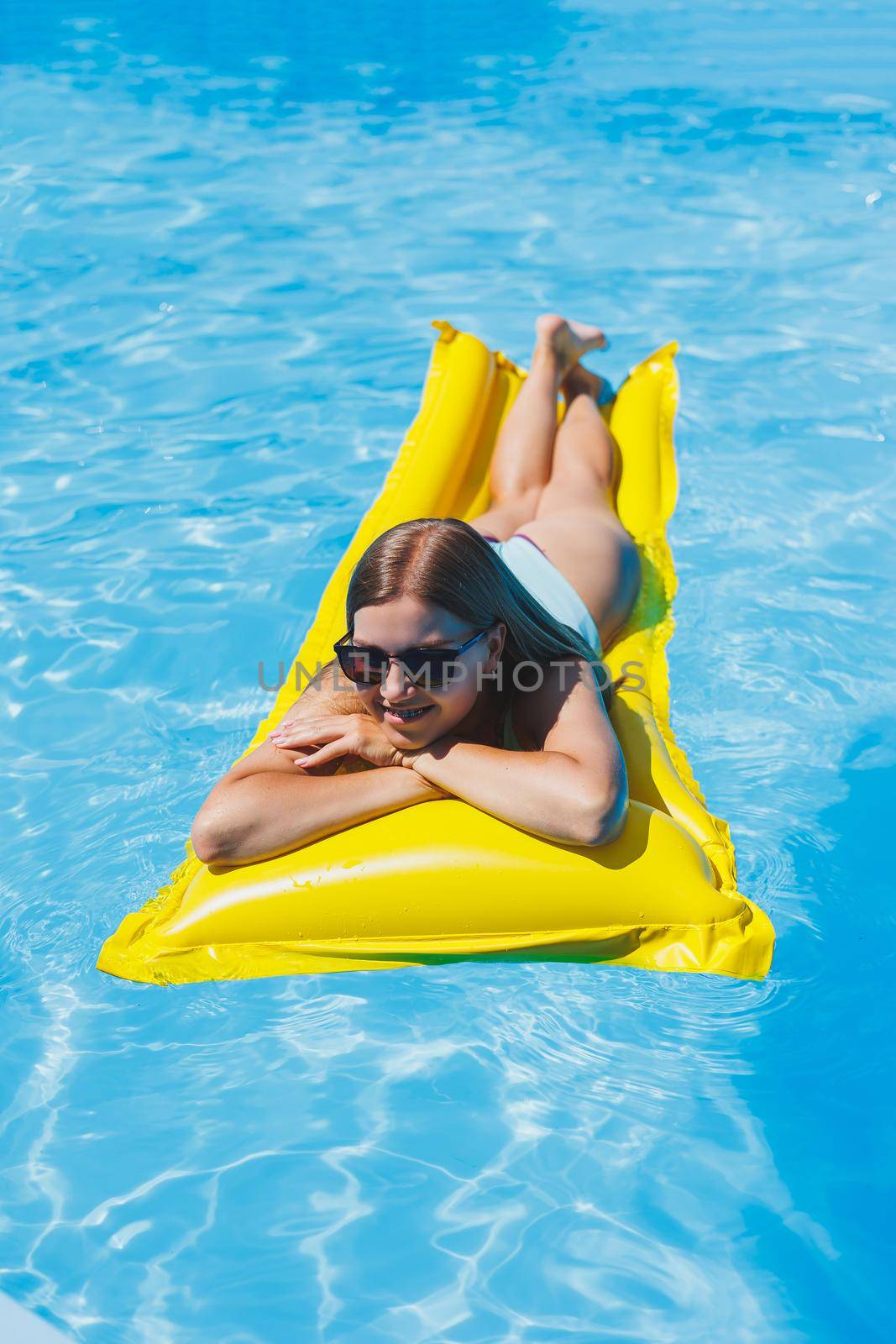 Pretty young woman in swimsuit and sunglasses lying on yellow inflatable mattresses in the pool. Enjoy sunbathing and relaxation.