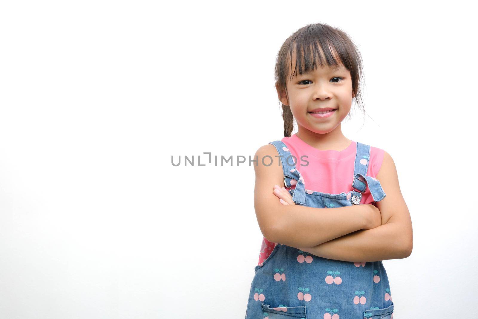 Asian cute little girl in jean overalls smiling showing her white teeth looking at the camera standing with her arms crossed on a white background with copy space.