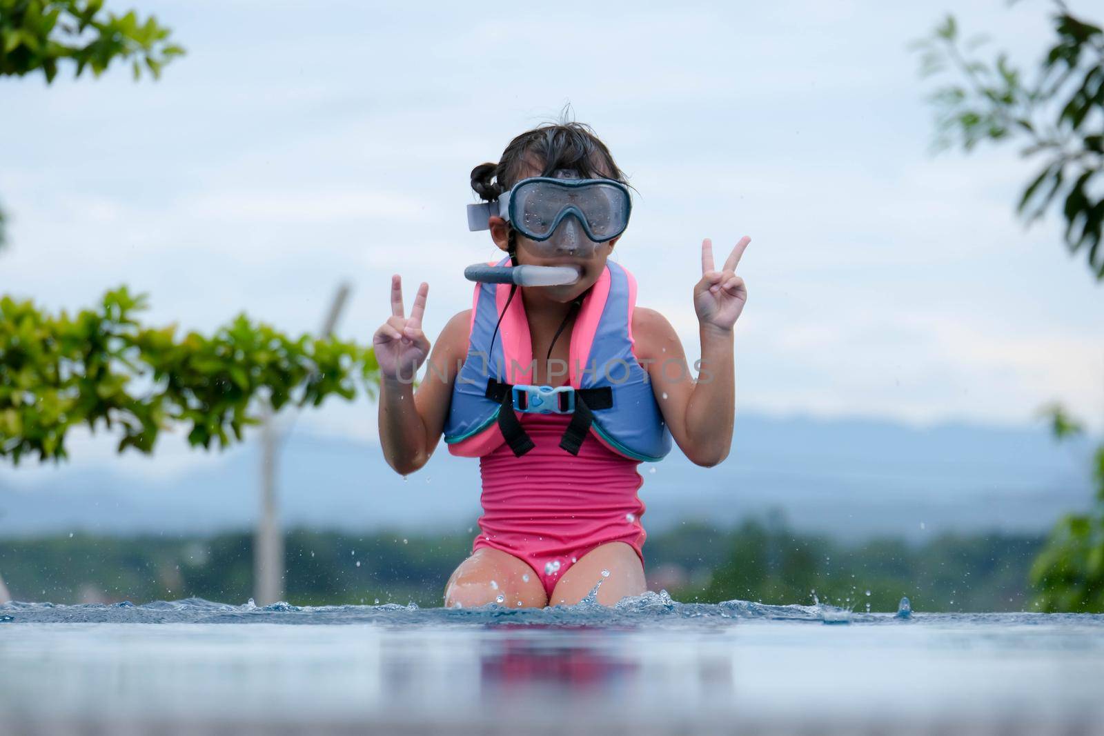 Cute little girl wearing goggles is sitting by the pool preparing for a diving lesson. Happy little girl is swimming and playing in outdoor swimming pool. Healthy Summer Activities for Kids. by TEERASAK