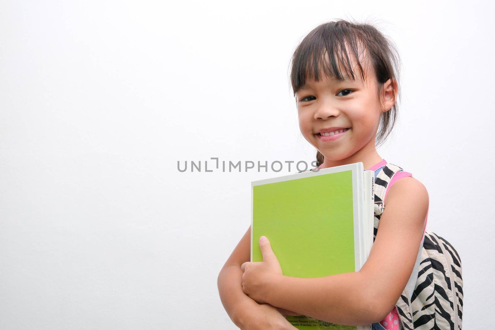 Back to school. Smiling little girl carrying a backpack holding books looking at the camera on a white background with copy space. Girl glad ready to study.