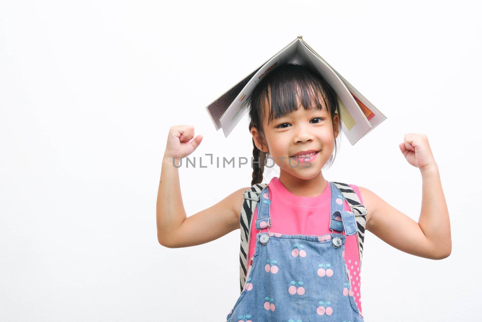 Back to school. Smiling little girl carrying a backpack holding books on her head looking at the camera on a white background with copy space. Girl glad ready to study. by TEERASAK