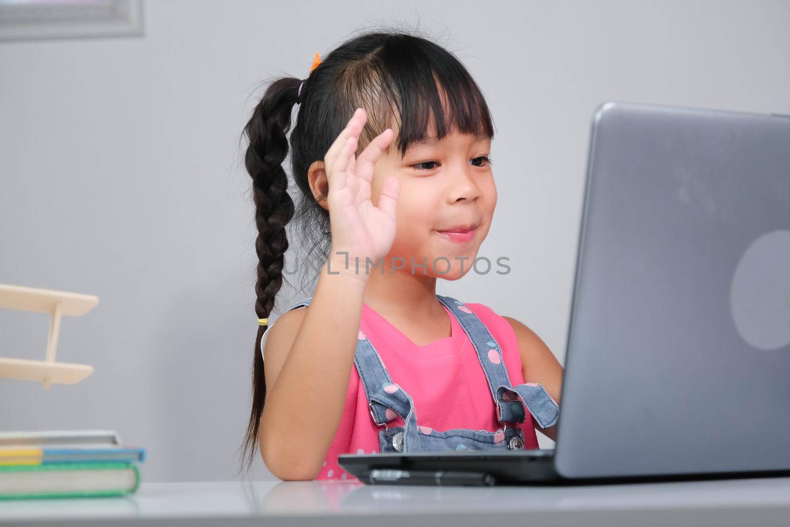 Smiling little Asian girl studying online having video call distant class with teacher using laptop. Happy little girl wave greeting with tutor while studying online with laptop at home. homeschooling by TEERASAK
