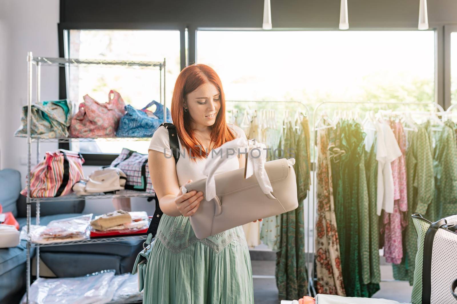 smiling young red-haired girl shopping in a small clothes shop looking at a handbag. girl spending time shopping. concept of shopping. natural light from the window of the window.