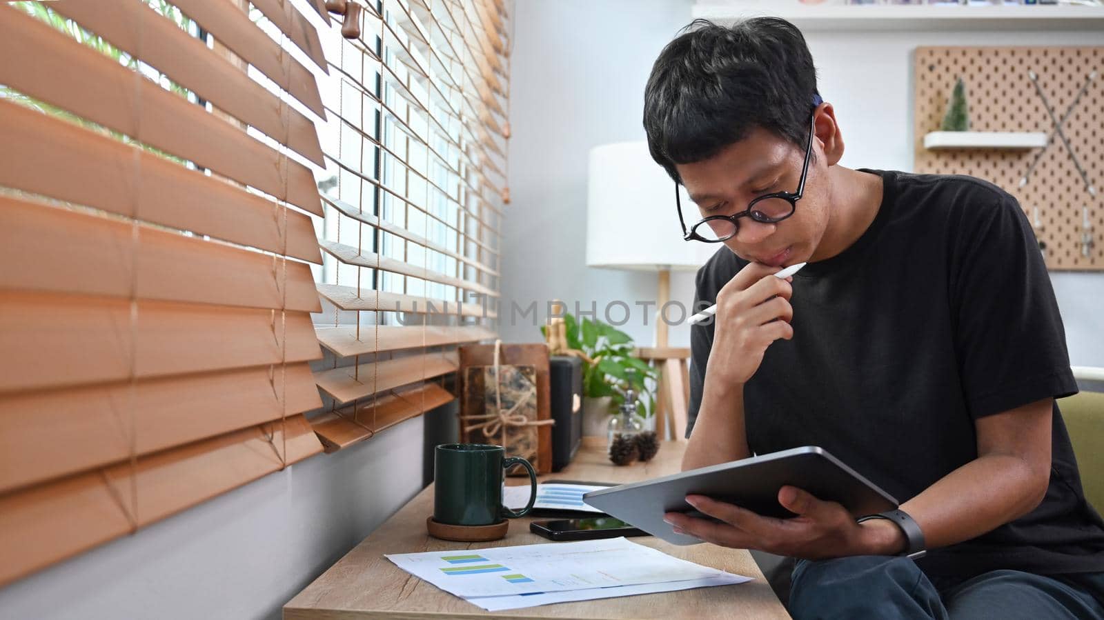 Thoughtful asian man analyzing data stock market on digital tablet at home. by prathanchorruangsak