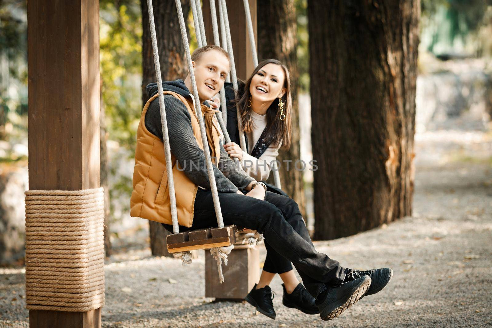 Side view of young cute couple riding on swing in autumn in warm clothes of basic colors in park outdoors by Rom4ek