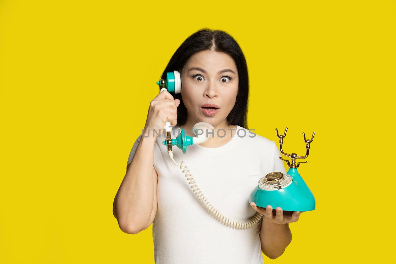 Shocked of winning lottery middle aged asian woman with vintage, retro telephone in hands excited, surprise face expression, isolated on yellow background. Communication concept.