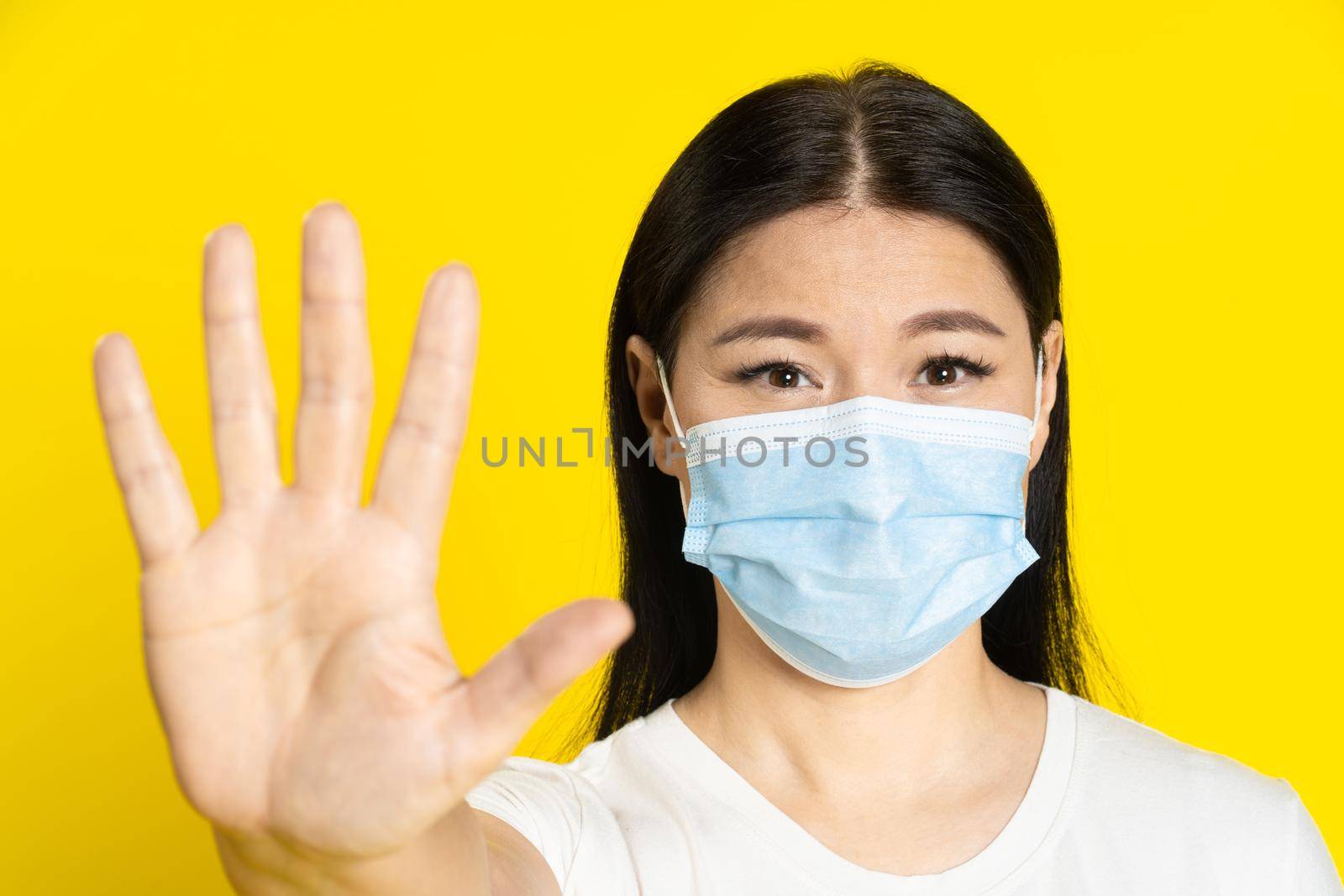 Asking to stop middle aged asian woman wearing medical mask gesturing to keep out with positive eyes on camera isolated on yellow background. Pandemic concept, monkey pox or coronavirus.