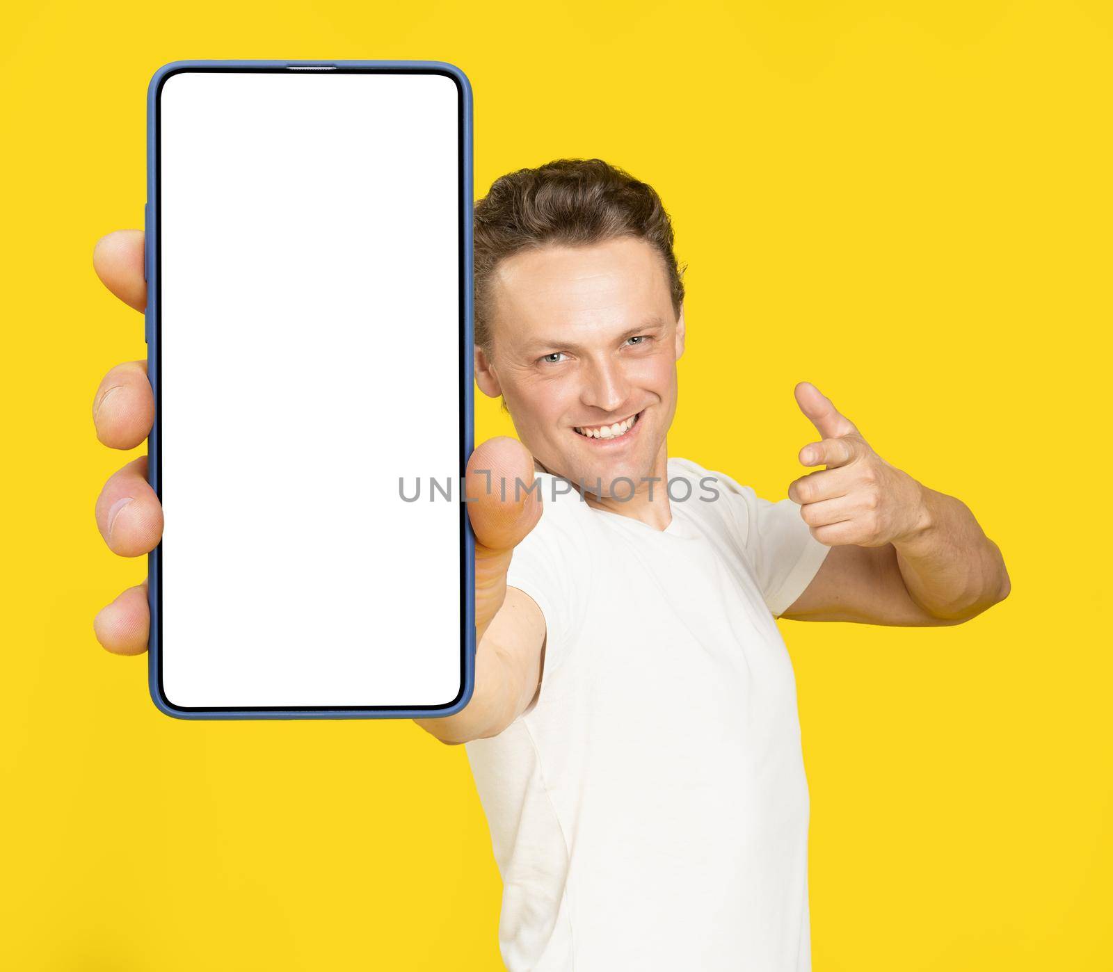 Holding huge smartphone with white screen handsome blond man pointing at white empty screen, wearing white t-shirt and jeans isolated on yellow background. Mobile app advertisement, great offer by LipikStockMedia