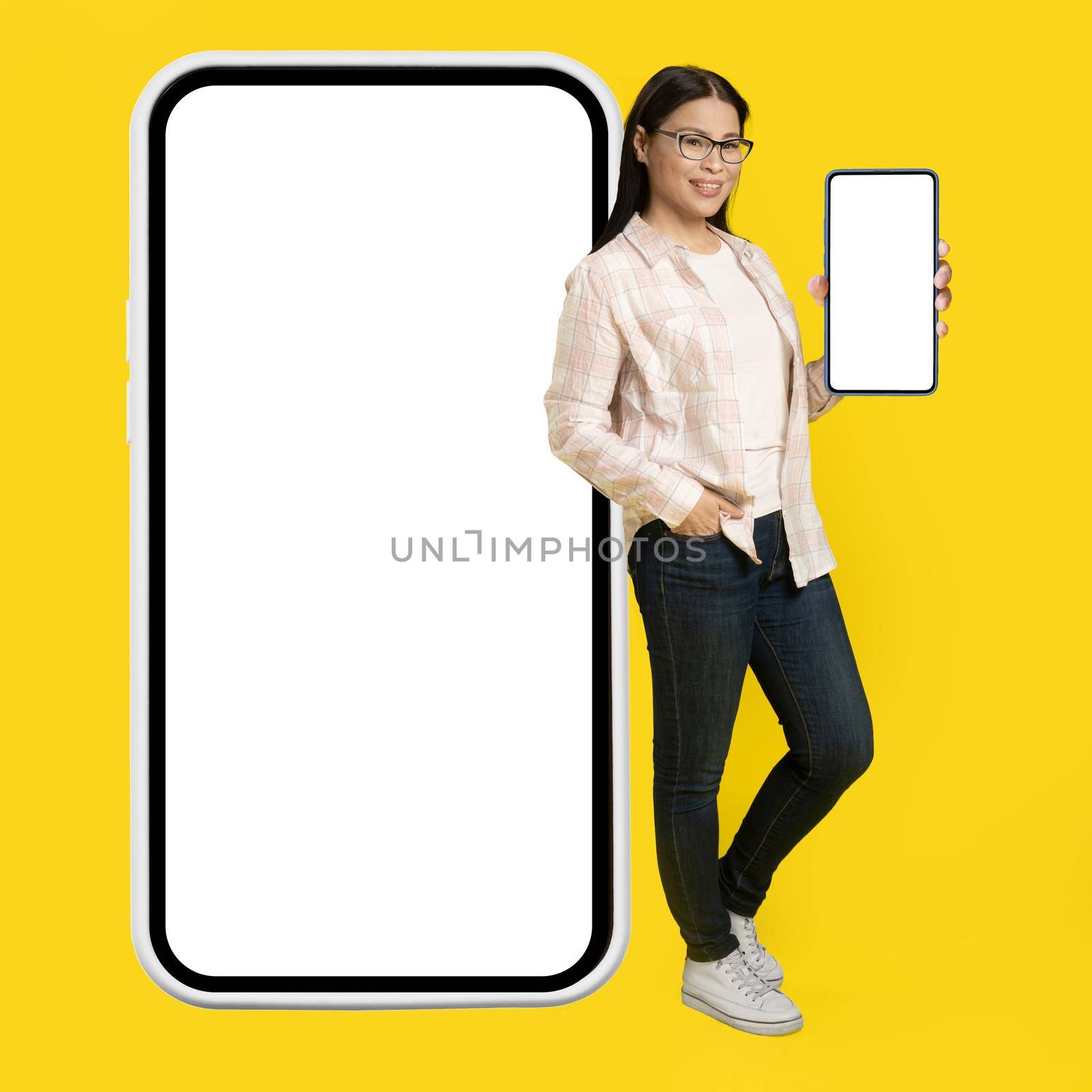 Showing white screen on phone charming middle aged asian woman leaned back on giant, huge smartphone with white screen wearing casual isolated on yellow background. Free space mock up by LipikStockMedia