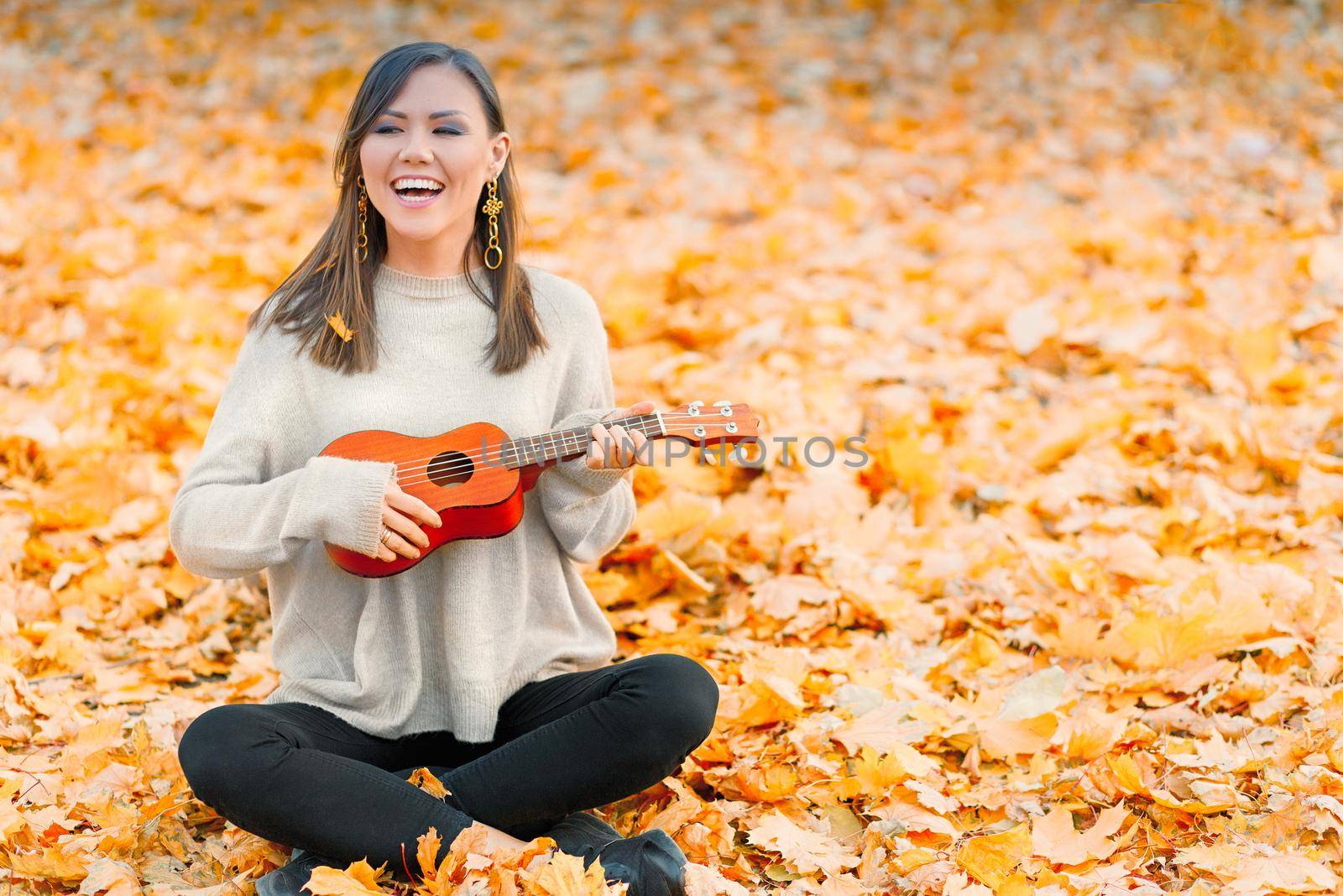 Young cheerful asian woman playing ukulele in autumn park on fallen leaves.