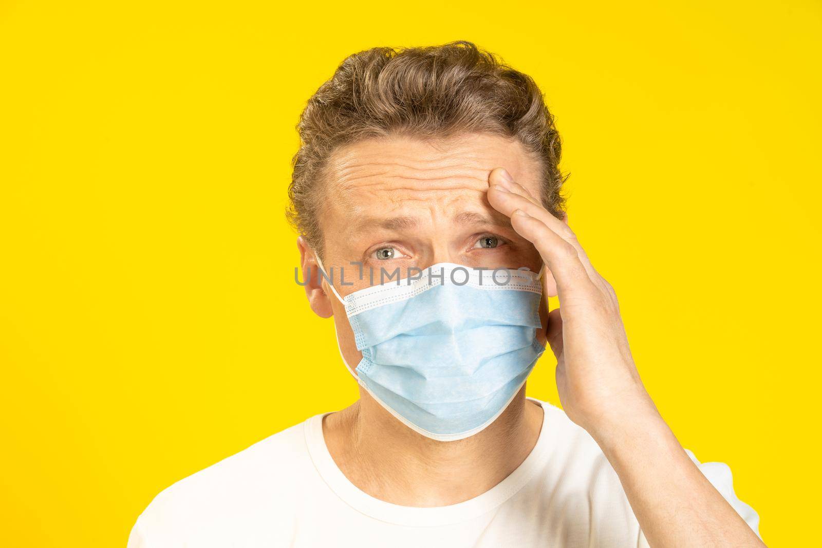 Young man suffering from a headache is holding his head wearing a medical mask on his face. Sick young man in face mask feel migraine touching his head. A sick caucasian man in protective face mask.