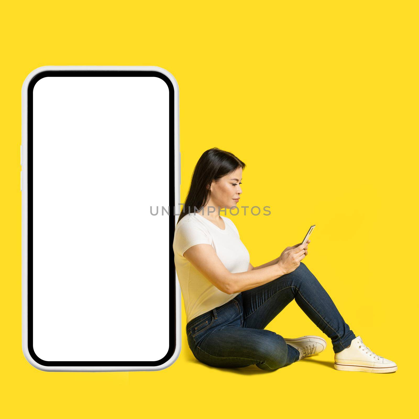 Sitting on the floor middle aged asian woman with phone in hands leaned on giant, huge smartphone with white screen wearing white t-shirt and jeans isolated on yellow background. Free space mock up by LipikStockMedia