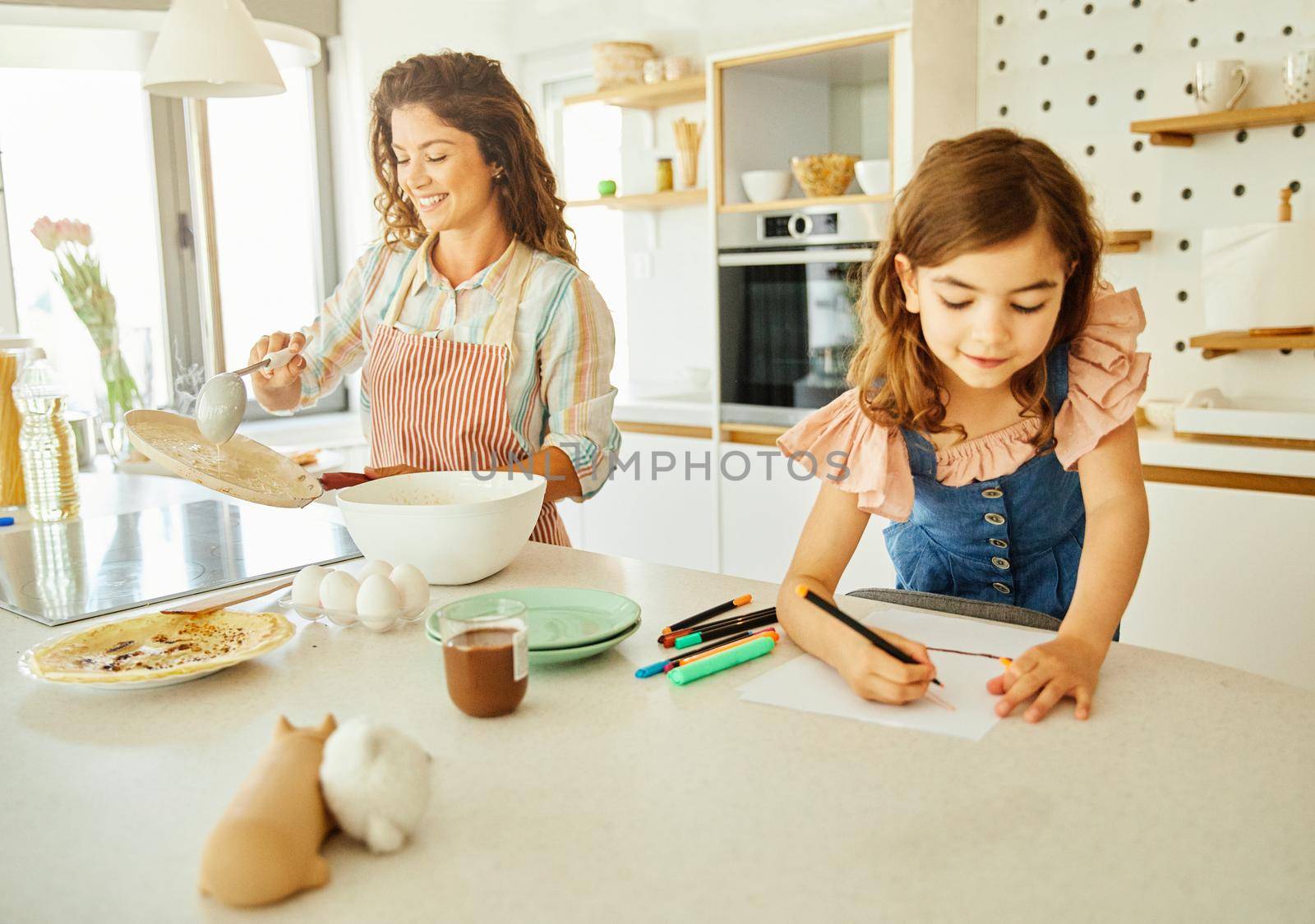 Mother preparing pancakes and daughter doing homework in the kitchen at home