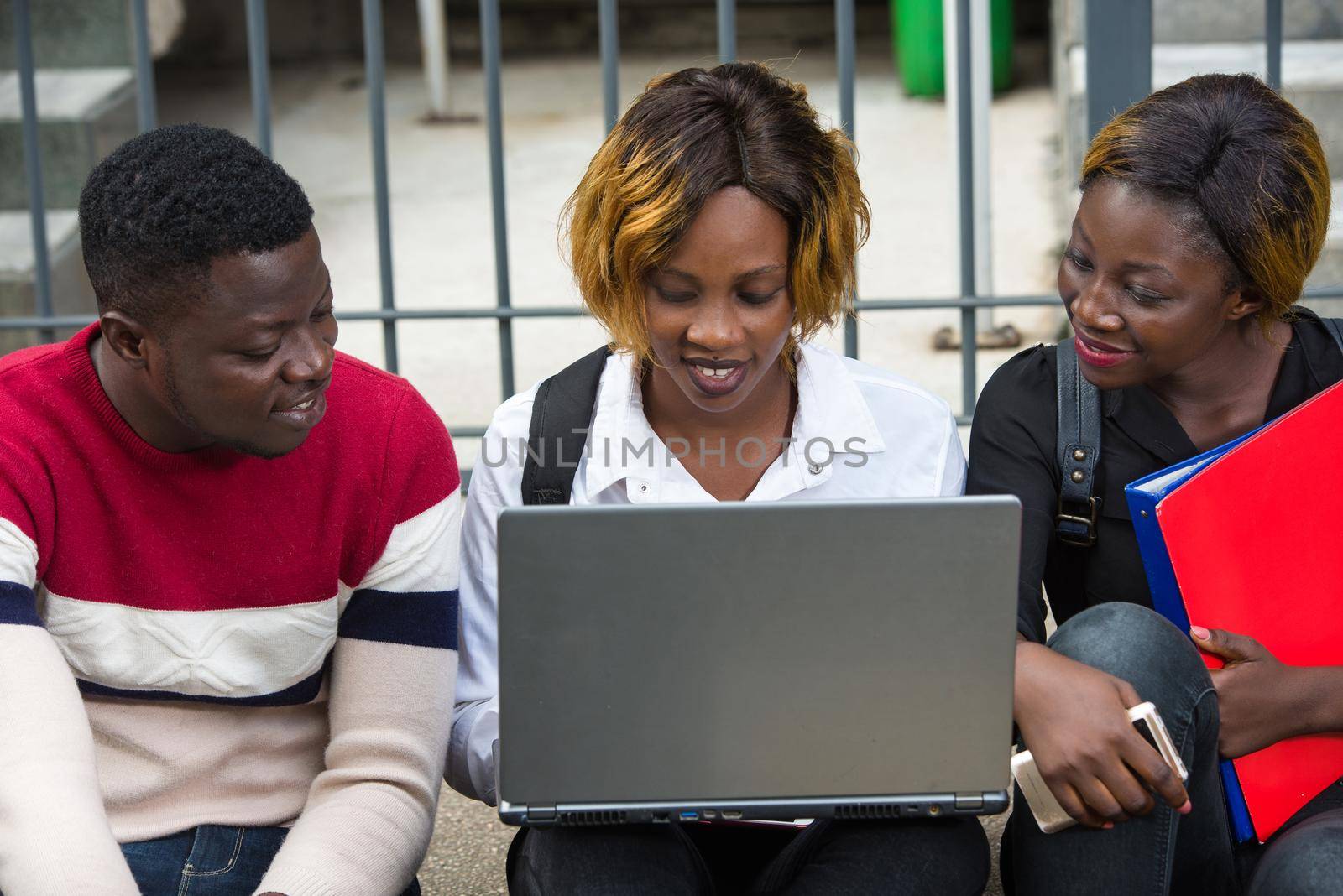 Group of attractive smiling young casual dressed students using laptop sitting outdoors on campus of university.