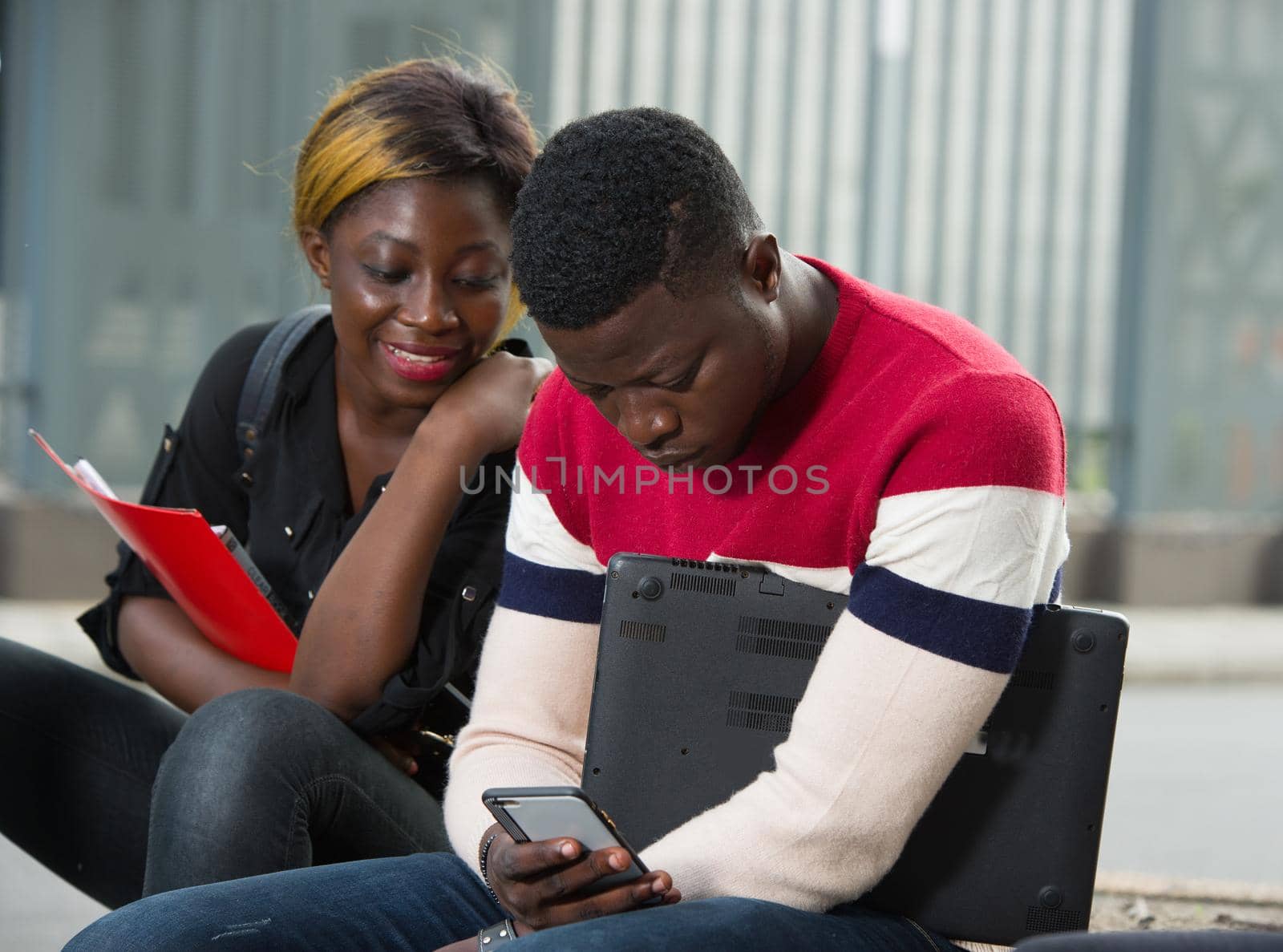 two young students outdoors studying with one cell phone, smiling girl sitting next to and looking at the mobile phone screen