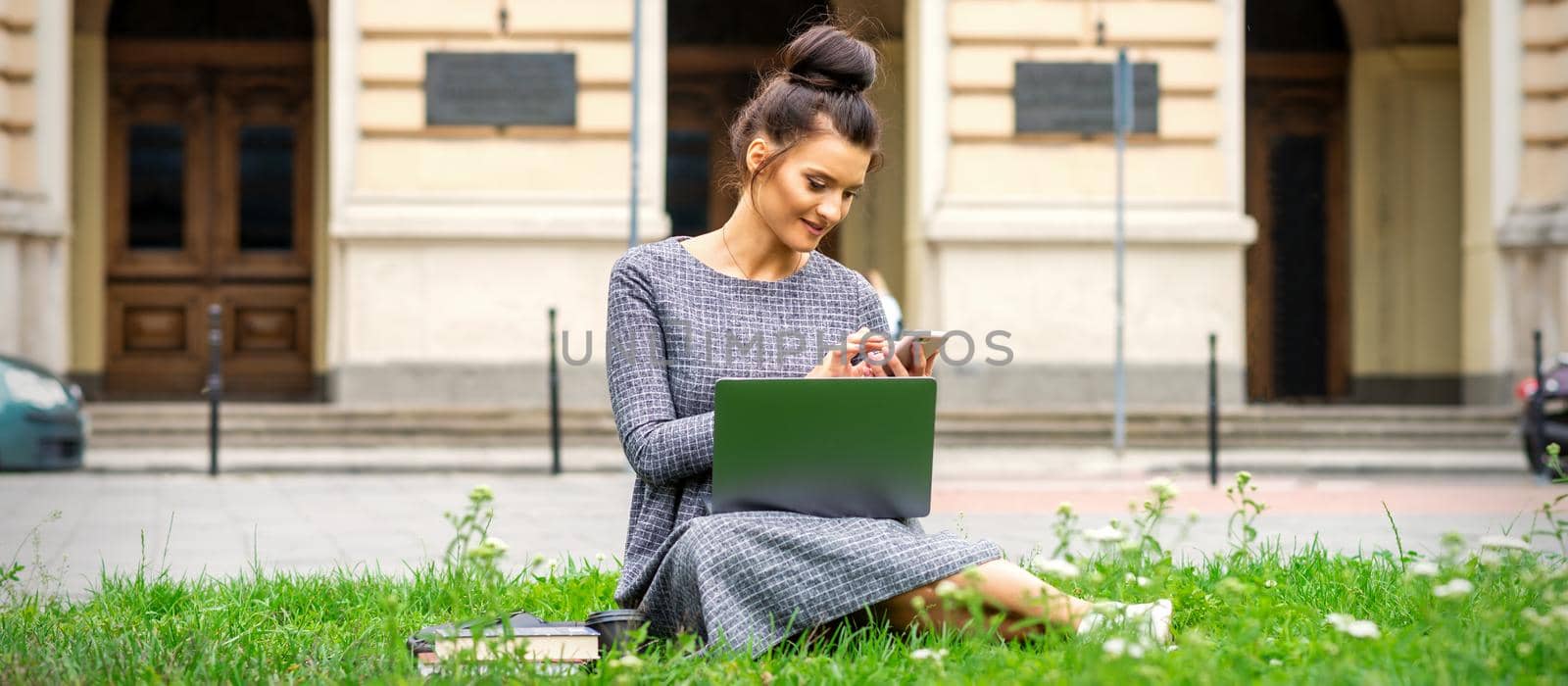 A young woman sits on the grass with a mobile phone and laptop computer against the background of a university