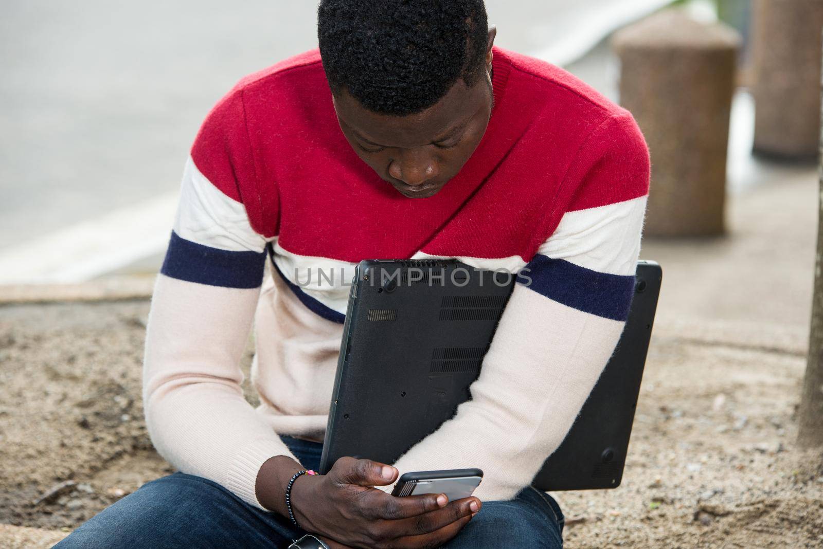 Pensive young student sitting outside, holding a laptop and looking on a cellphone,