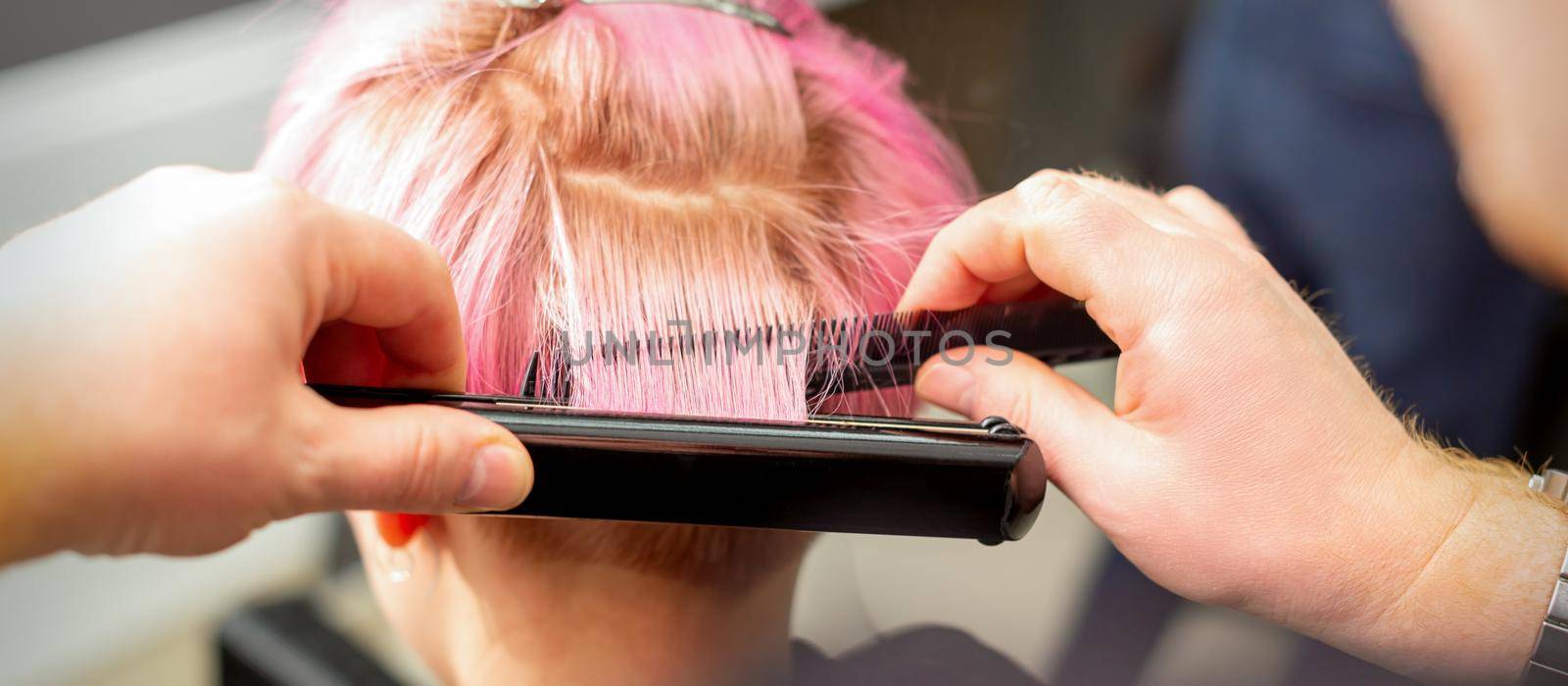 Close up back view of hairdresser's hands straightening short pink hair with a hair iron straightener in a hair salon