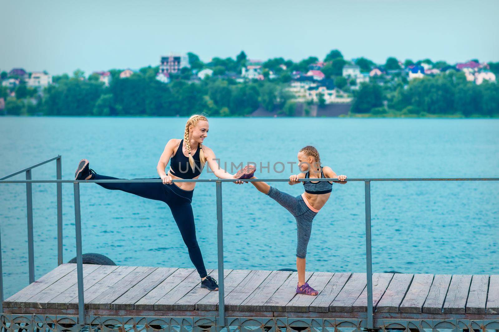 Woman and child are doing sport exercises stretching their legs on the pier the lake